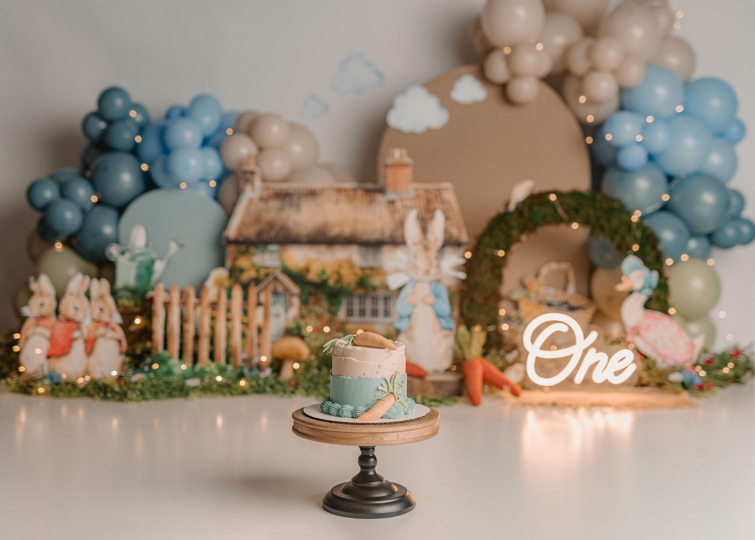 Peter Rabbit themed first birthday cake smash setup with blue balloons, fairy lights, and a decorative village backdrop.
