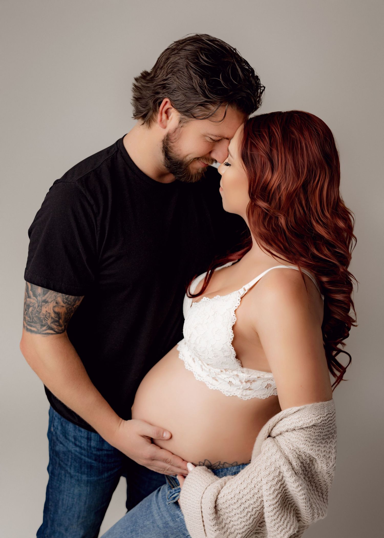 Bearded man in a black tee touches foreheads with his red-haired pregnant partner in a lace top while cradling her bare baby bump.