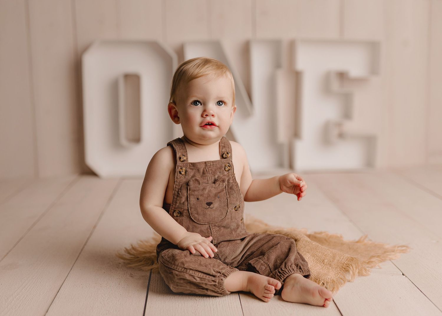 A baby in a brown dress sits and plays on a light wooden floor in a bright, airy room.