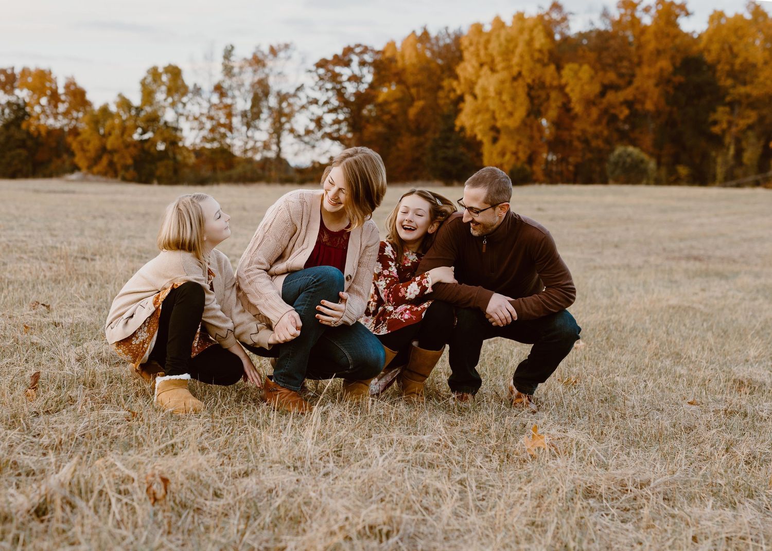 Family of four laughing together in an autumn field surrounded by golden trees during a fall outdoor photo session.