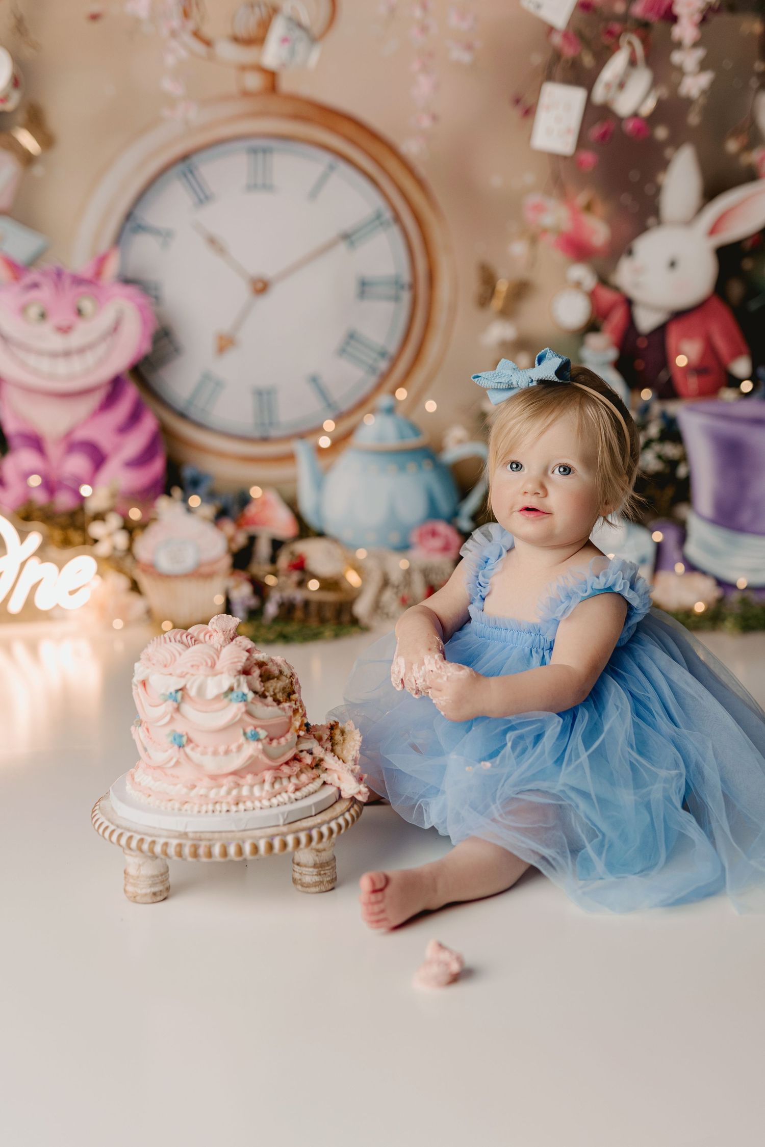 Baby girl in blue dress celebrates first birthday with Alice in Wonderland themed cake smash photo session. Lapeer cake smash session