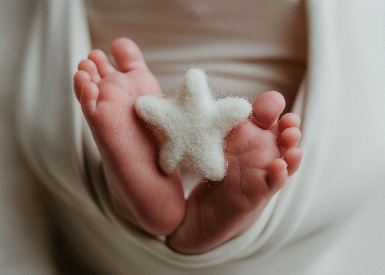 Close-up of newborn baby feet holding a white felt starfish, wrapped in a soft cream blanket.