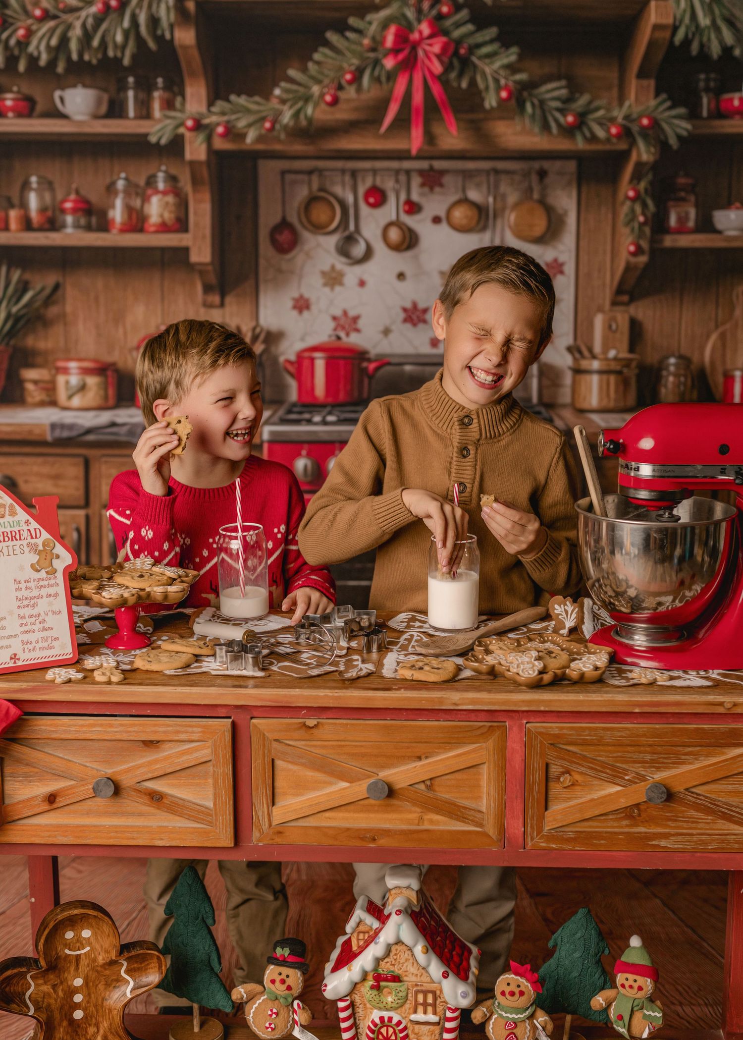 Children sit at a festive wooden counter decorated with gingerbread houses and Christmas ornaments in a cozy kitchen setting.