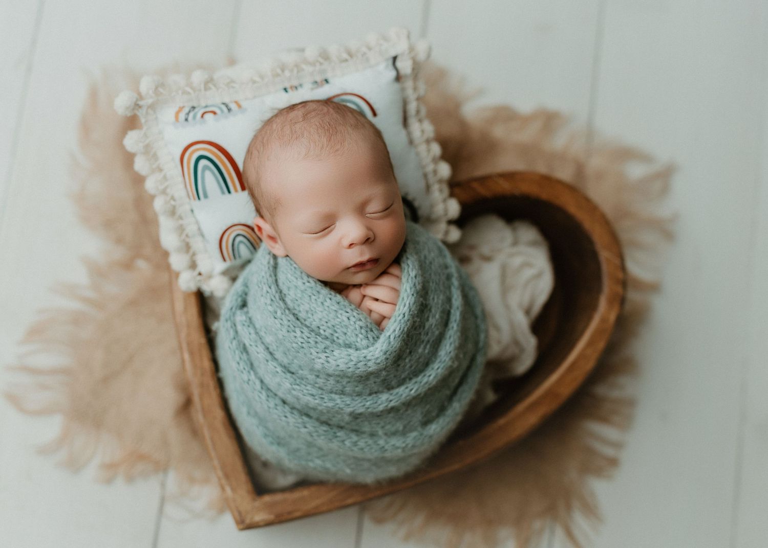 Newborn baby swaddled in sage green wrap sleeping in a wooden heart-shaped bowl with a rainbow pillow.