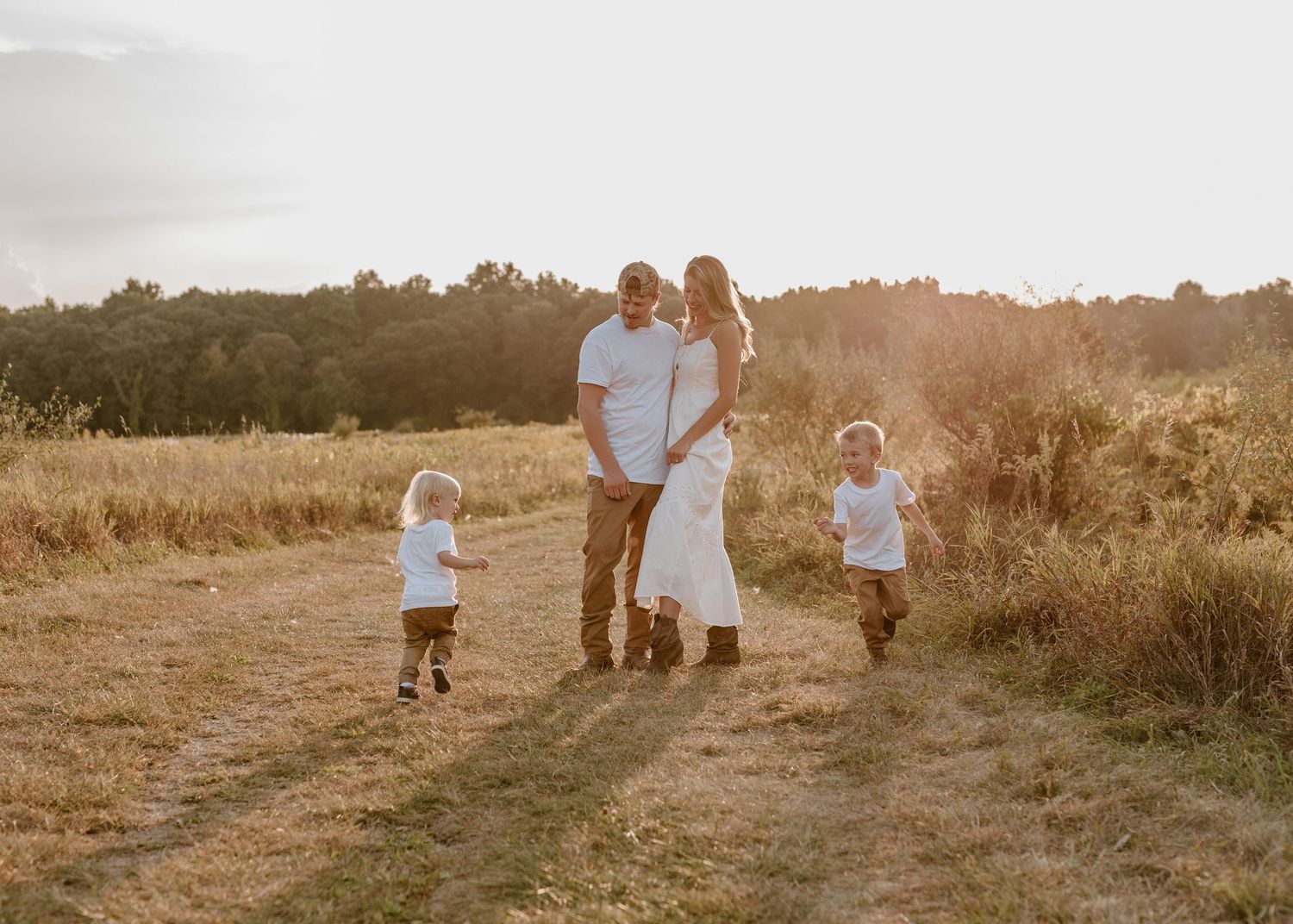 Family of four walking on a sunlit field path at golden hour, parents and two toddlers in white shirts.