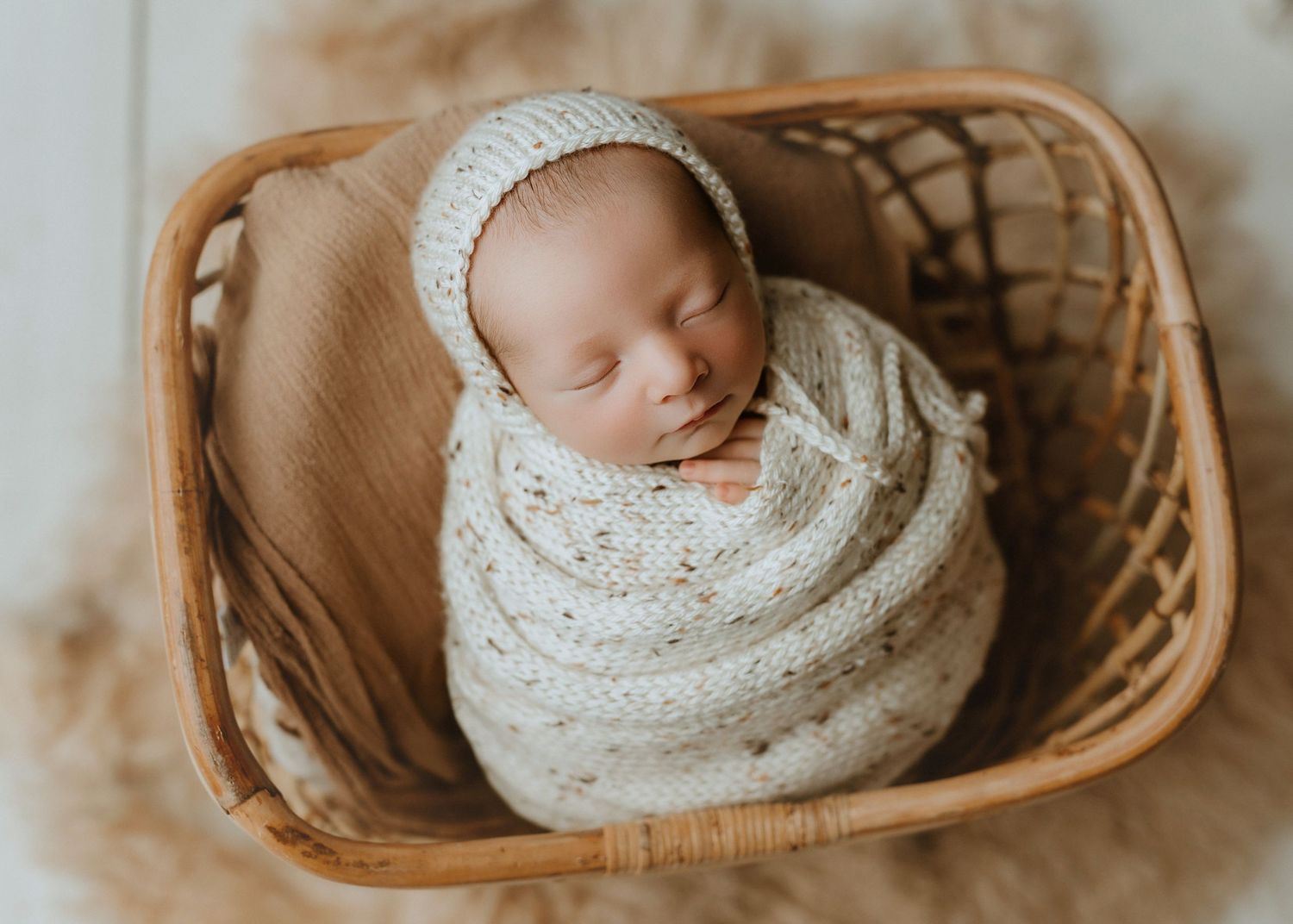 Newborn baby swaddled in cream knit wrap and bonnet, sleeping peacefully in a wicker basket.