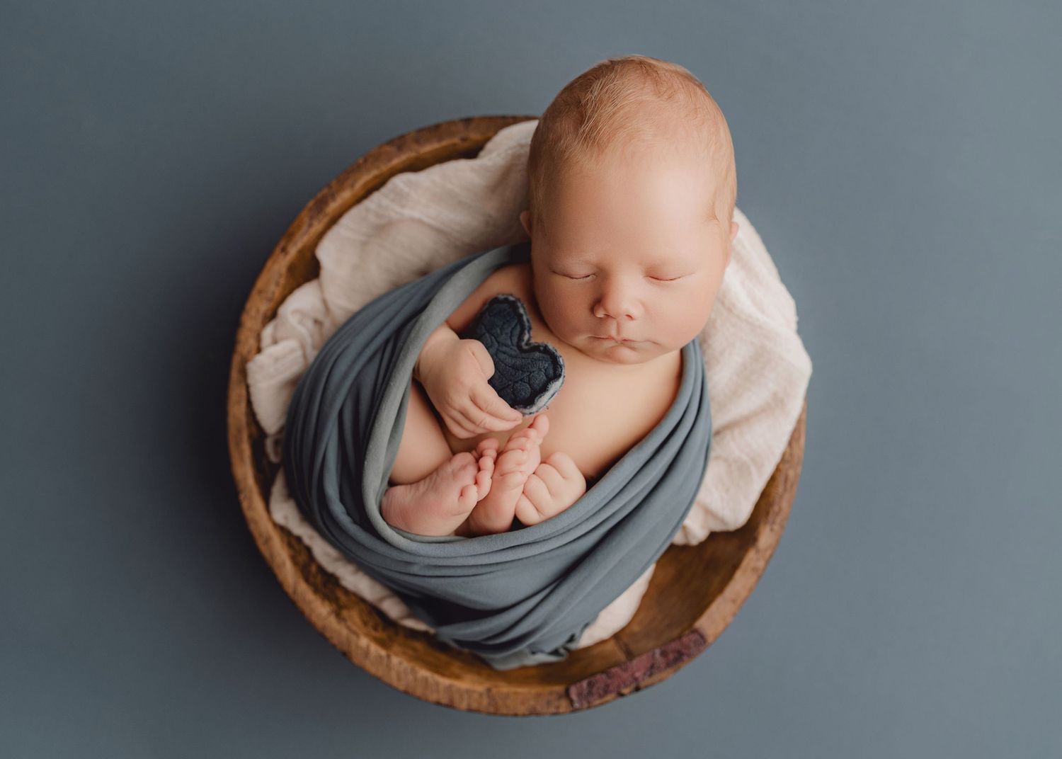 Peaceful newborn baby wrapped in soft gray fabric sleeping soundly in rustic wooden bowl against neutral backdrop.
