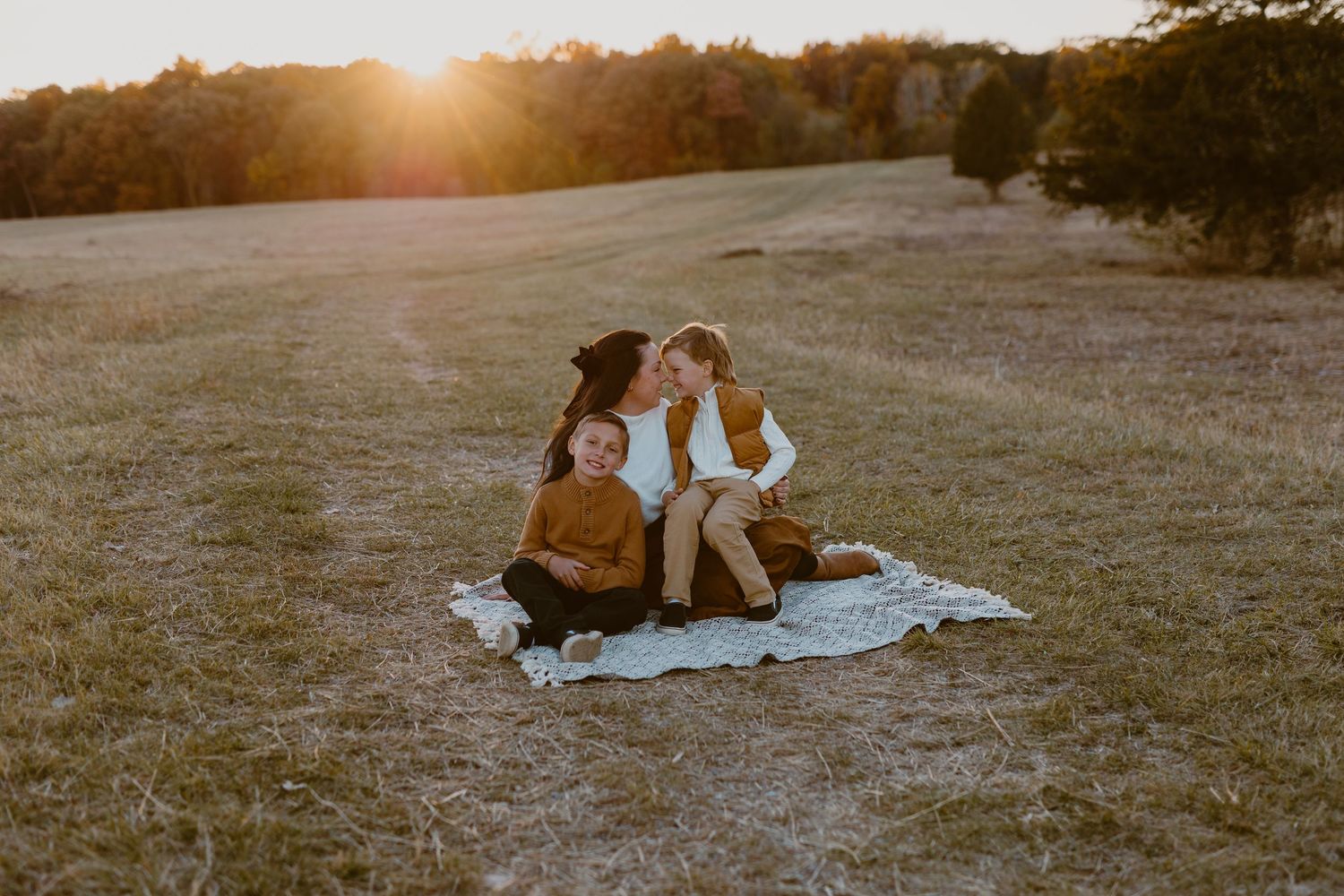 Family sitting on blanket in golden field at sunset, laughing together in warm autumn light.