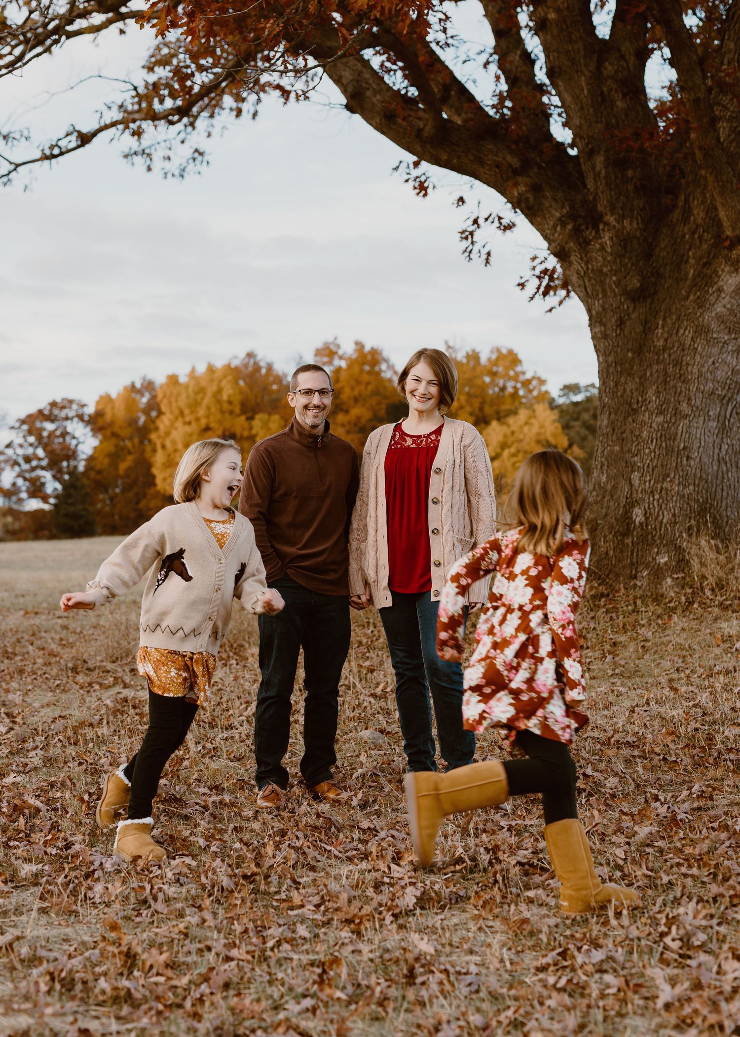 Family of four enjoying autumn outdoors, two children running toward smiling parents near a large oak tree with fall foliage.