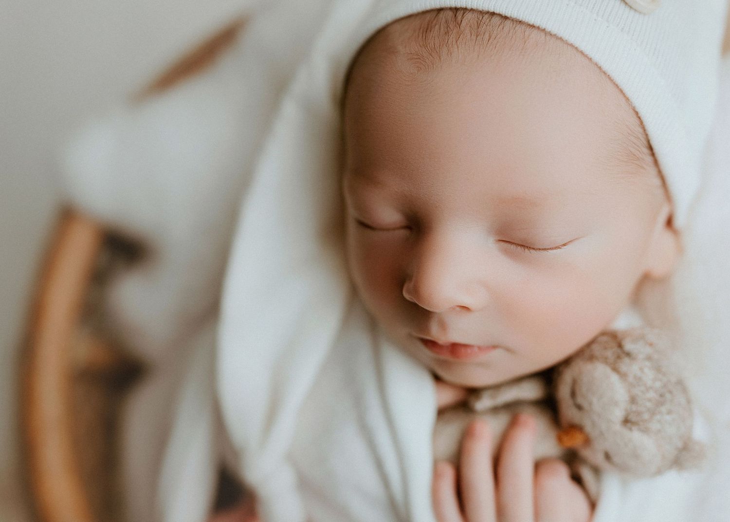 Peaceful newborn baby sleeping wrapped in white swaddle, holding tiny teddy bear, soft natural light photography.