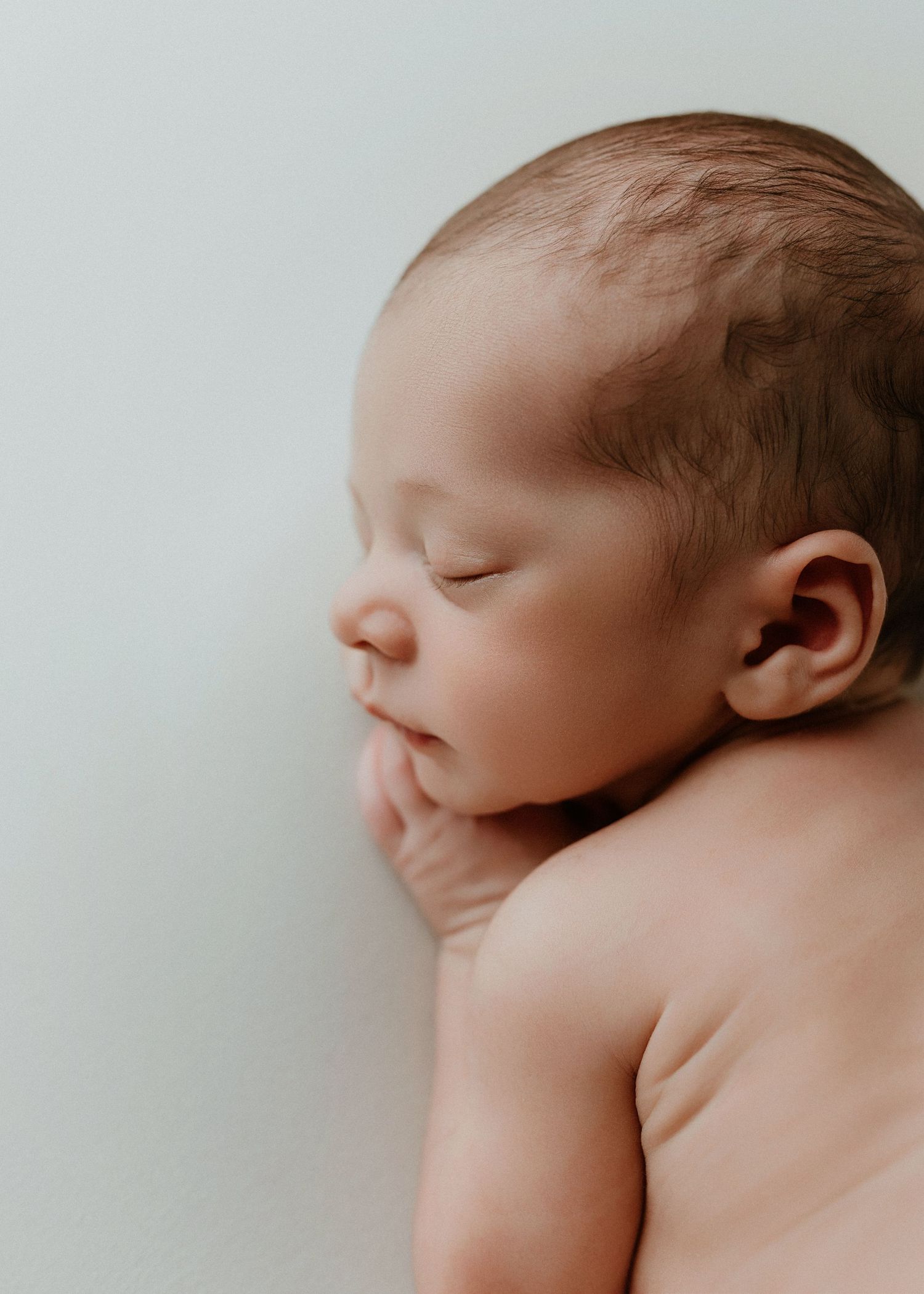 Peaceful sleeping newborn baby with soft brown hair resting on hands against a light background.