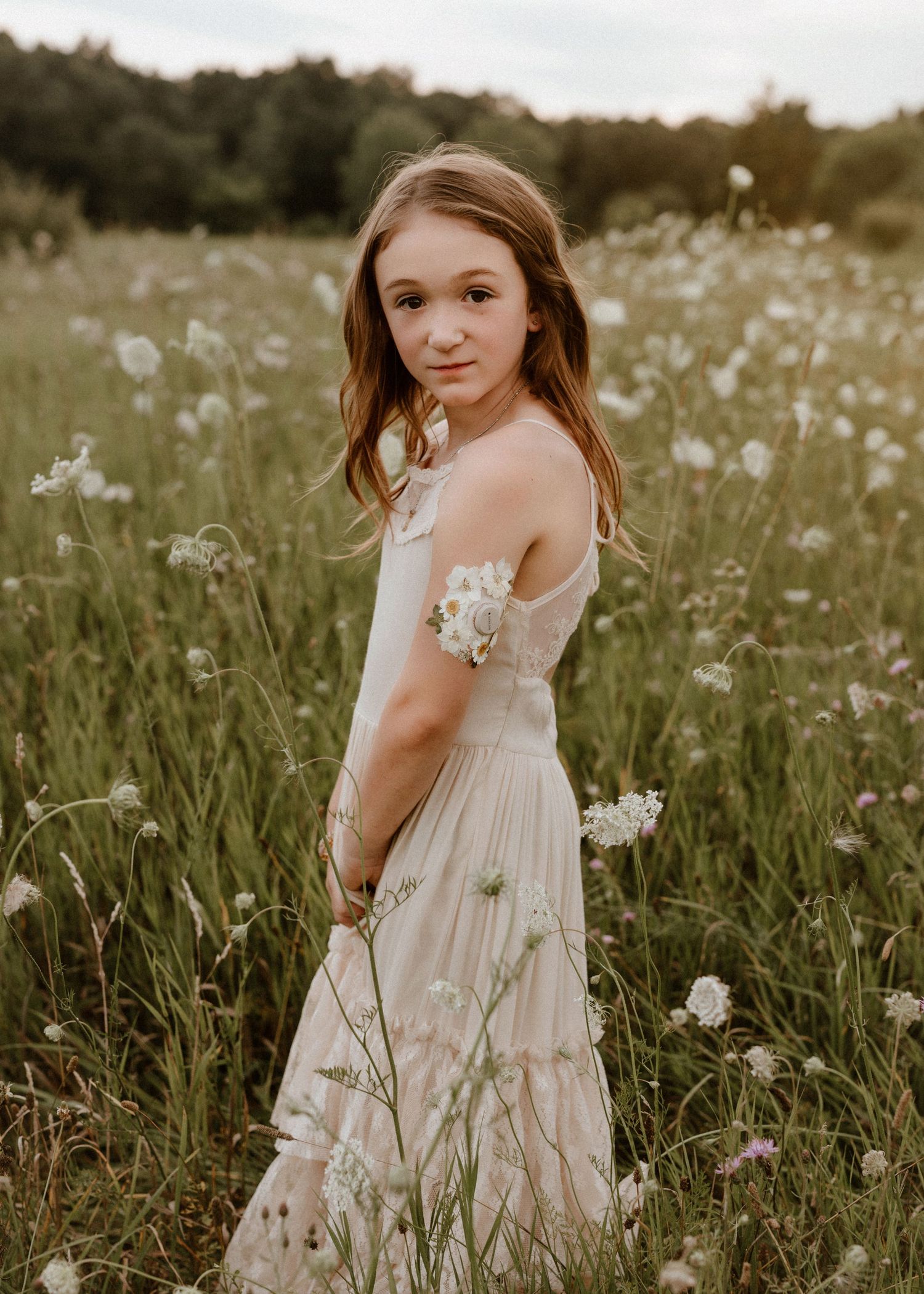 Young girl in white dress standing in wildflower field, looking over shoulder, golden hour light.