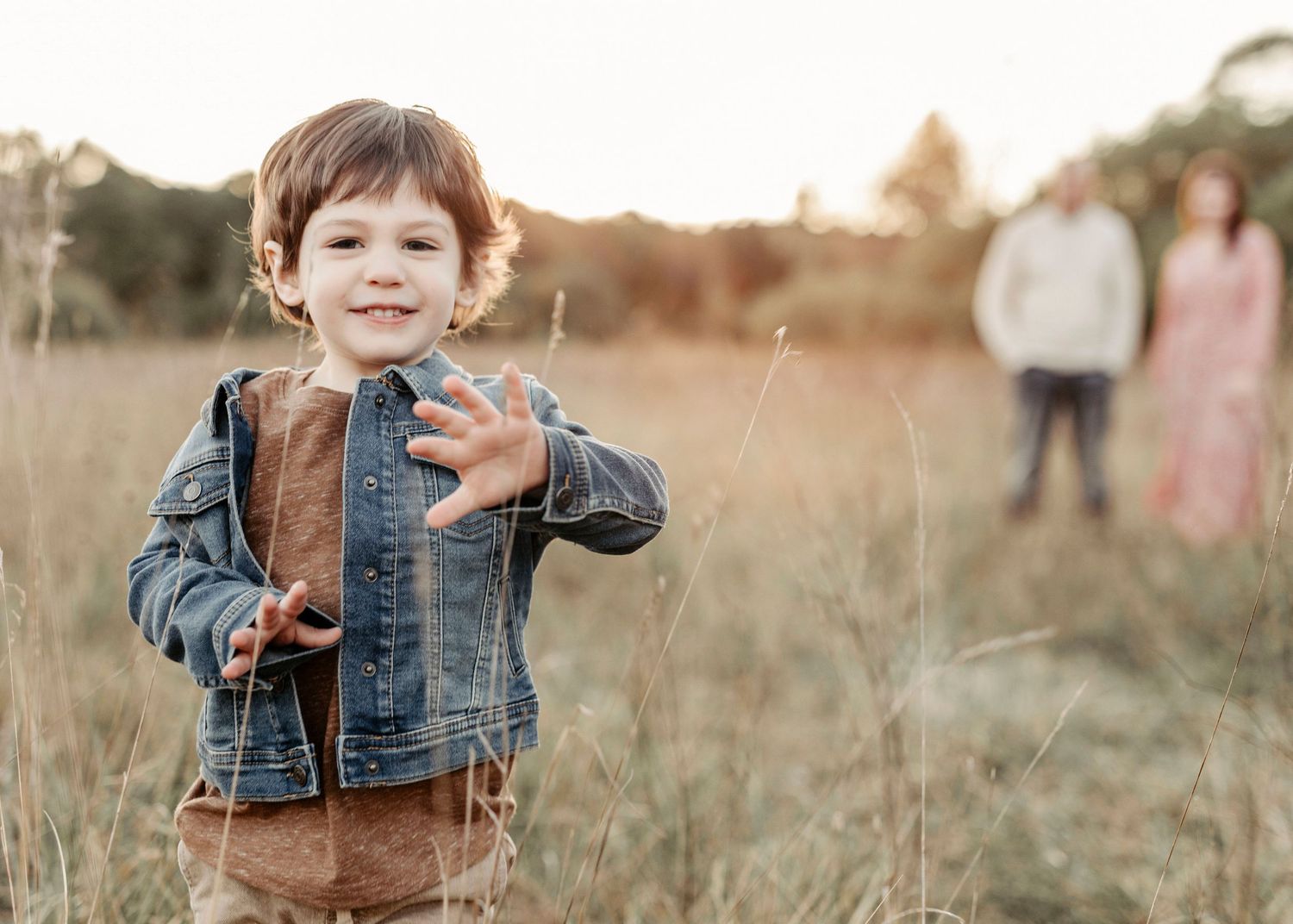 A smiling toddler boy in a denim jacket reaches toward the camera in a golden field with parents standing behind.