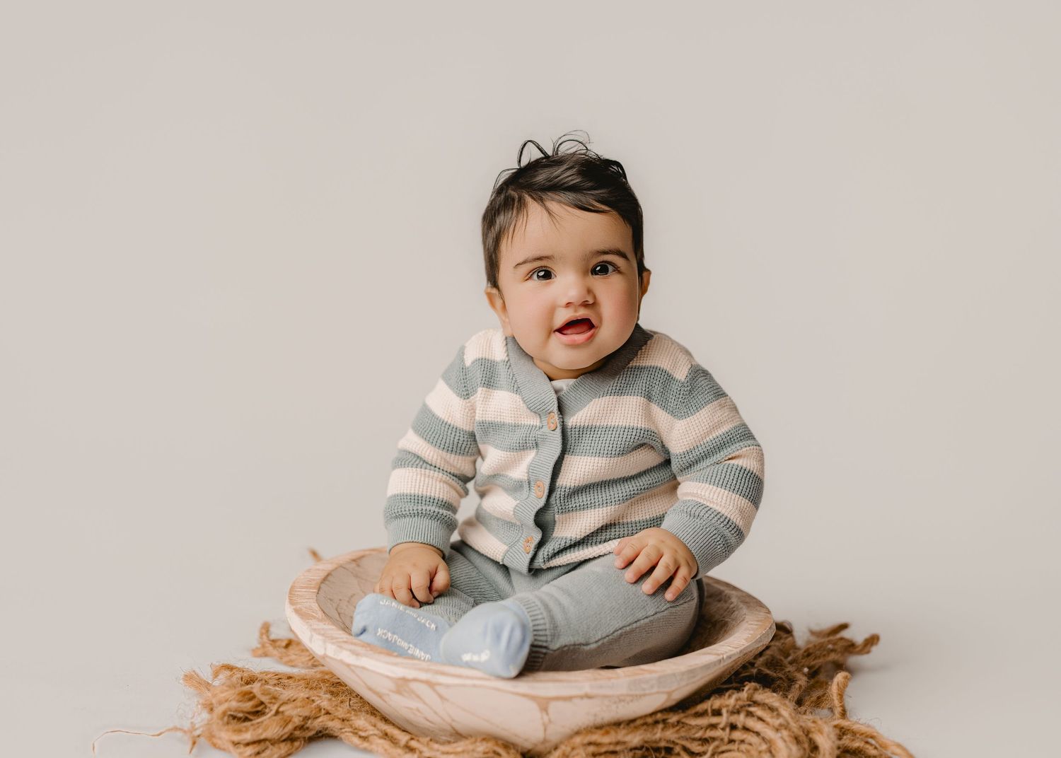 An adorable baby in a striped cardigan sits in a wooden bowl on a neutral background.