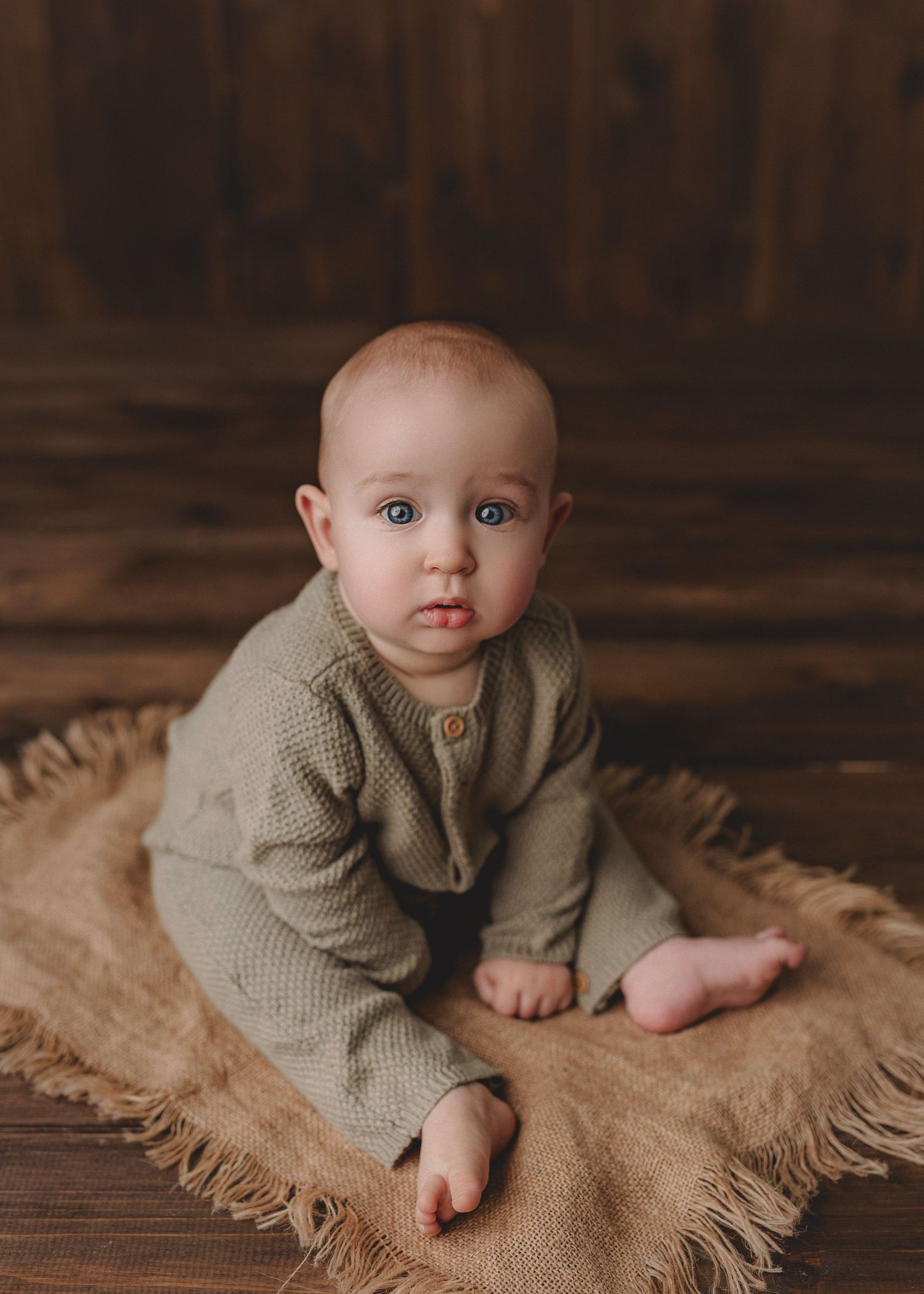 A baby with blue eyes sits on a burlap blanket wearing a sage green knit romper on a rustic wood floor.