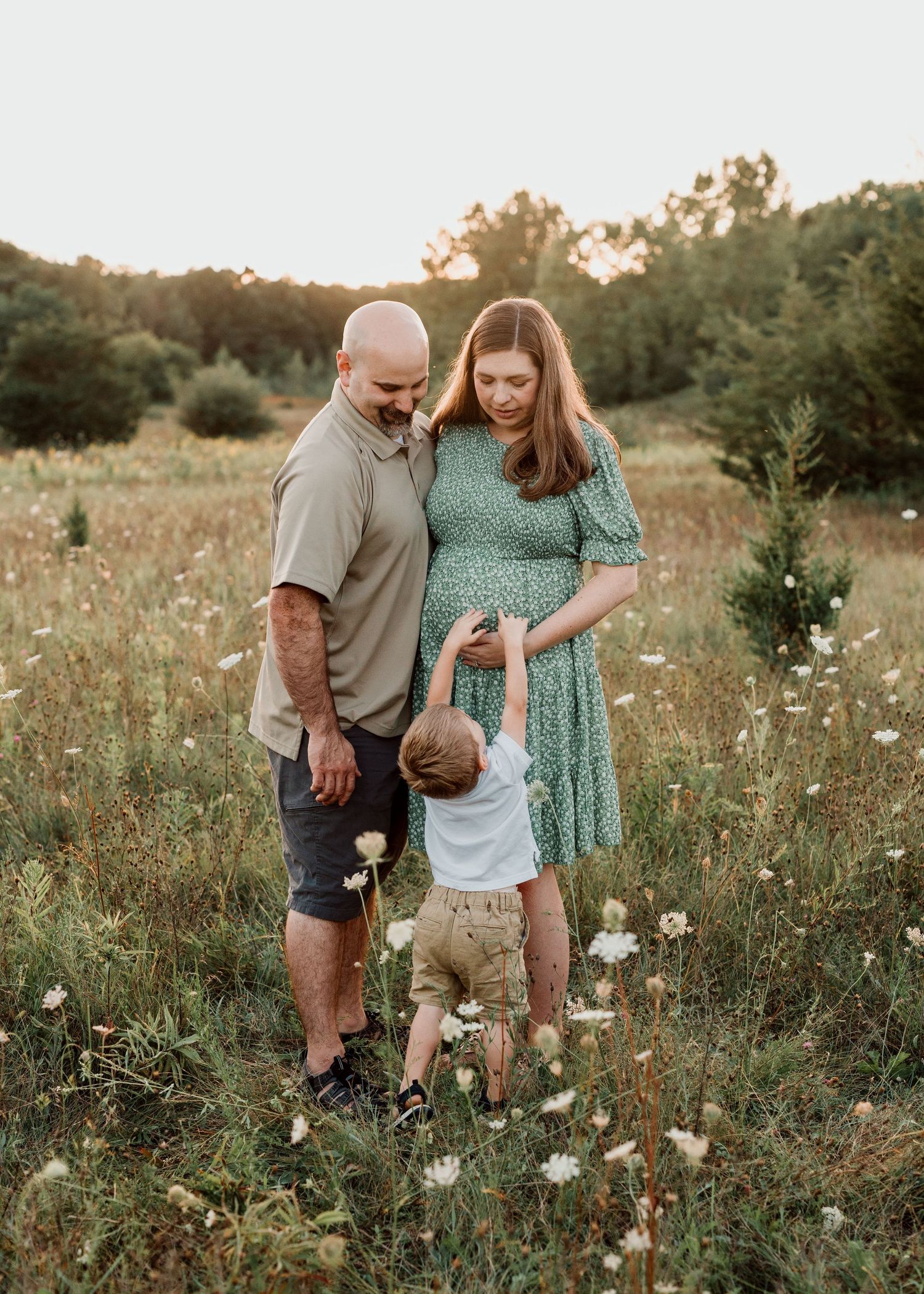 A pregnant woman and bald man watch their toddler in a sunny wildflower field at golden hour.