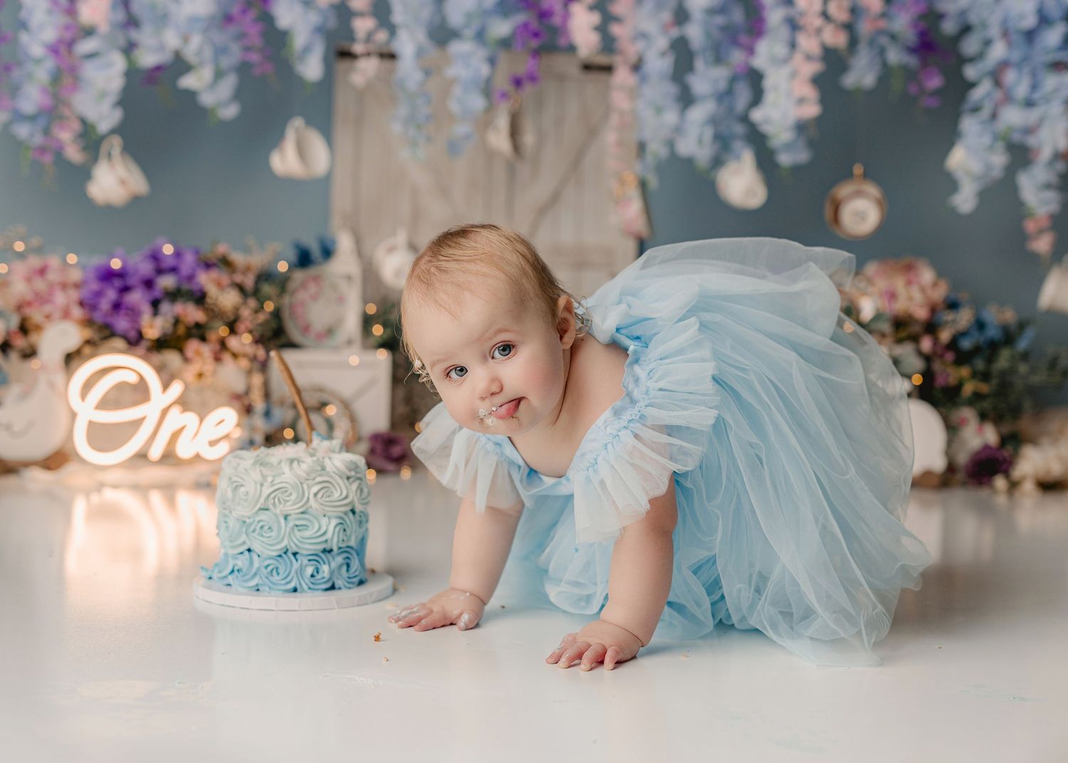 Baby girl in blue tutu dress crawls toward a blue rosette smash cake beside a glowing One sign at her first birthday.