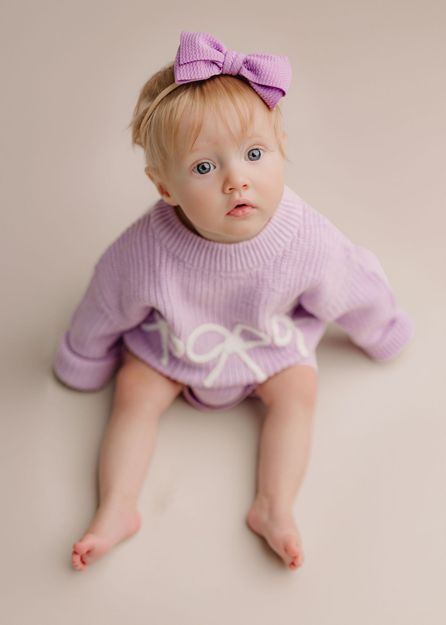 Baby girl in purple knit sweater and bow headband sits on neutral background looking up at camera.