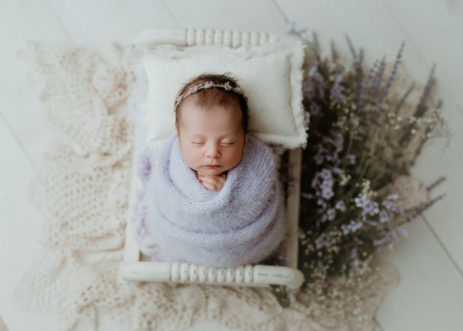 Newborn baby wrapped in lavender swaddle sleeping in tiny white wooden bed with floral headband and purple flowers.