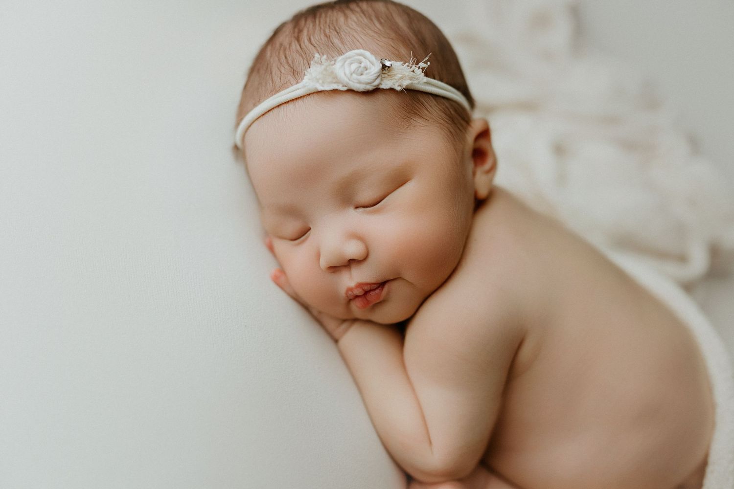 Peaceful sleeping newborn baby wearing a delicate white floral headband, posed on a soft cream background.