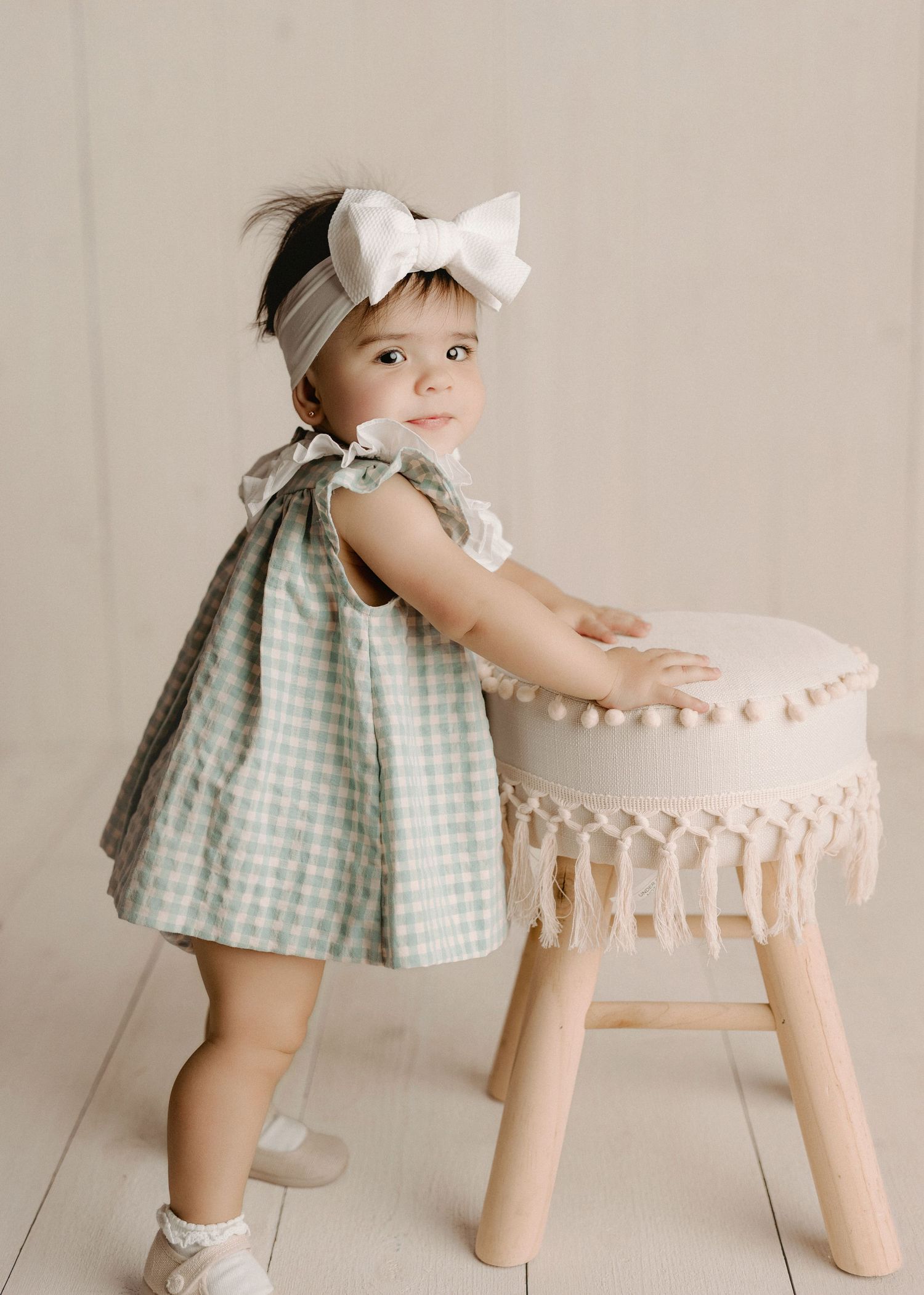 Adorable baby girl in sage green gingham dress with white bow headband standing next to a boho fringe stool.