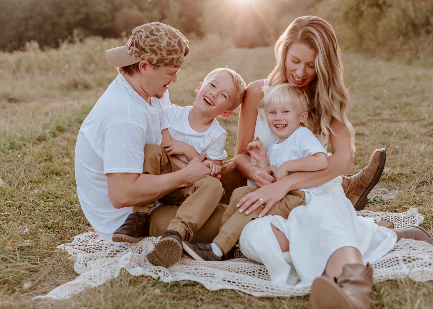 A happy family of four in white outfits laughing together on a blanket in a sunny field.