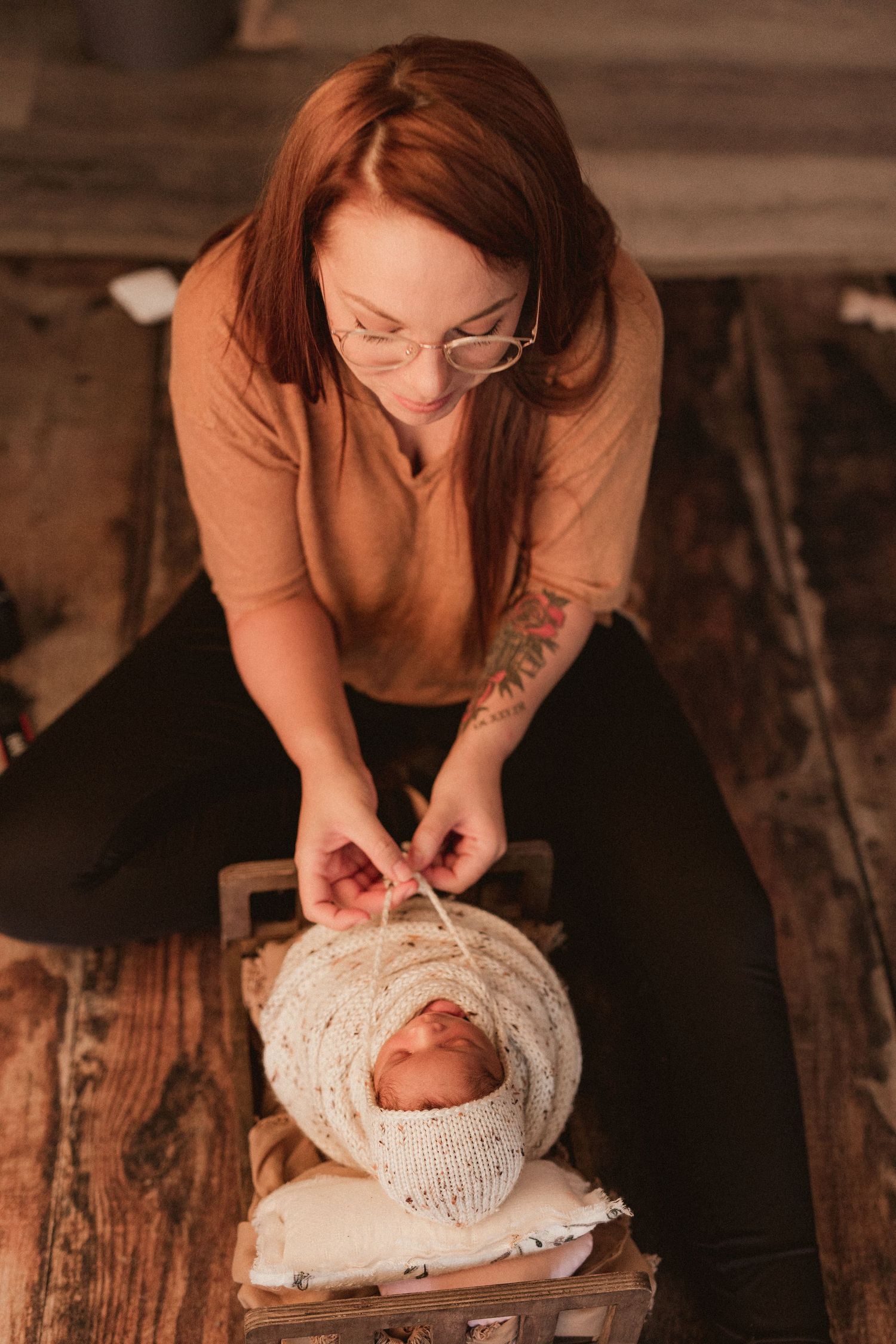 Professional photographer adjusts wrapping on sleeping newborn during rustic wooden floor photoshoot session. Oxford michigan