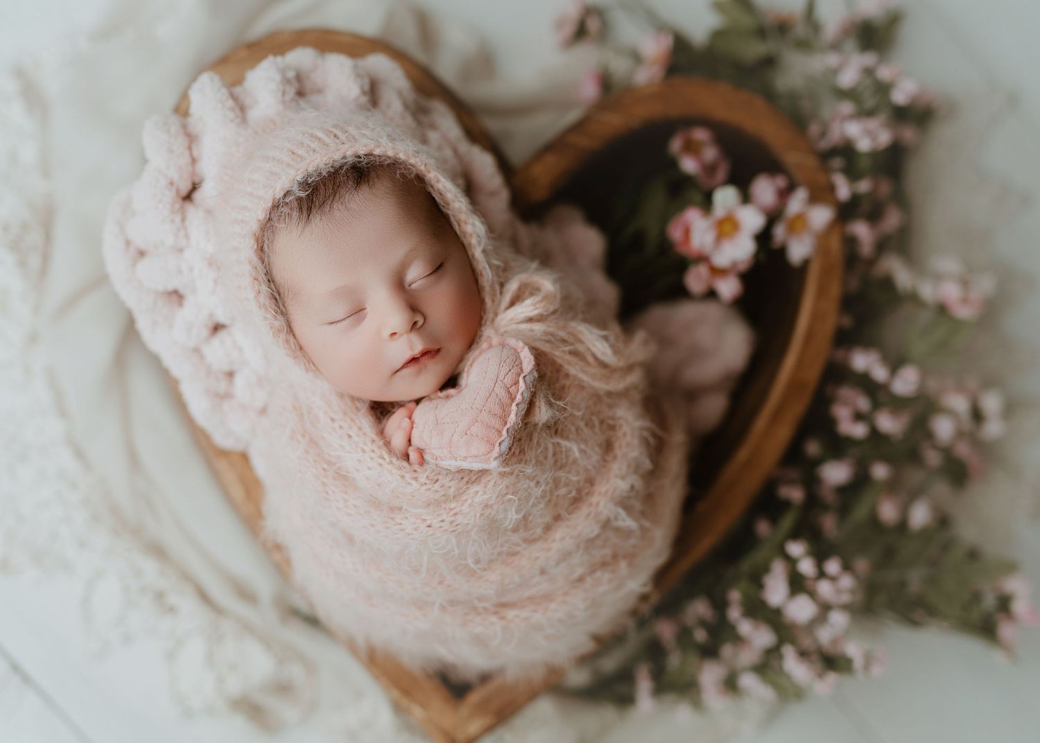 Newborn baby wrapped in pink knit swaddle wearing bonnet, sleeping in heart-shaped wooden bowl with flowers.
