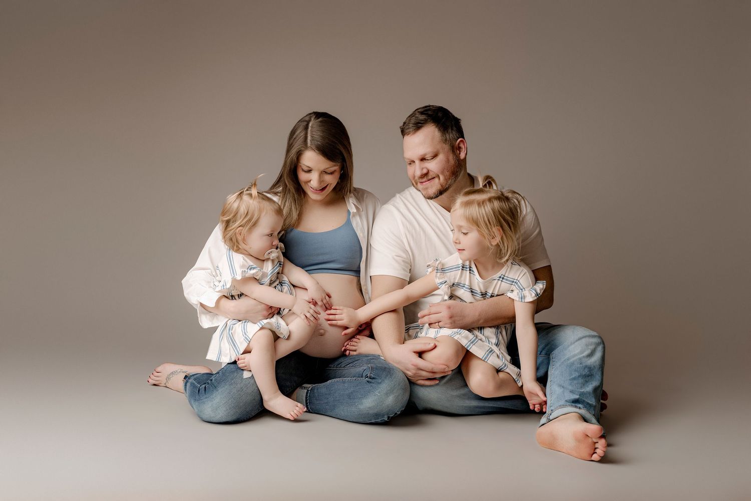 Smiling parents sit on the floor with two toddler daughters in a cozy maternity family portrait, mother cradling her baby bump.