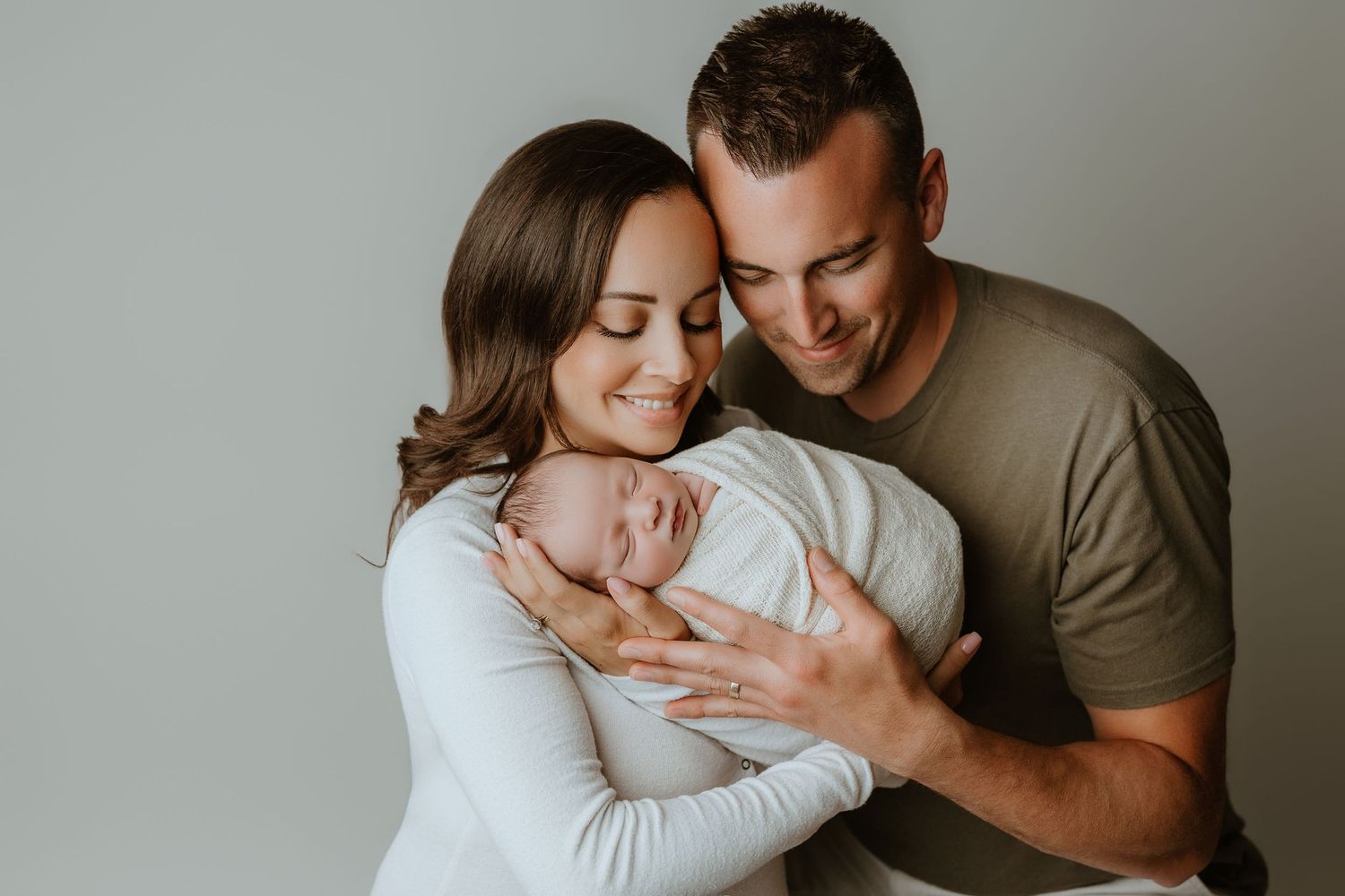 A happy couple smiling and cradling their newborn baby wrapped in white against a soft gray background.
