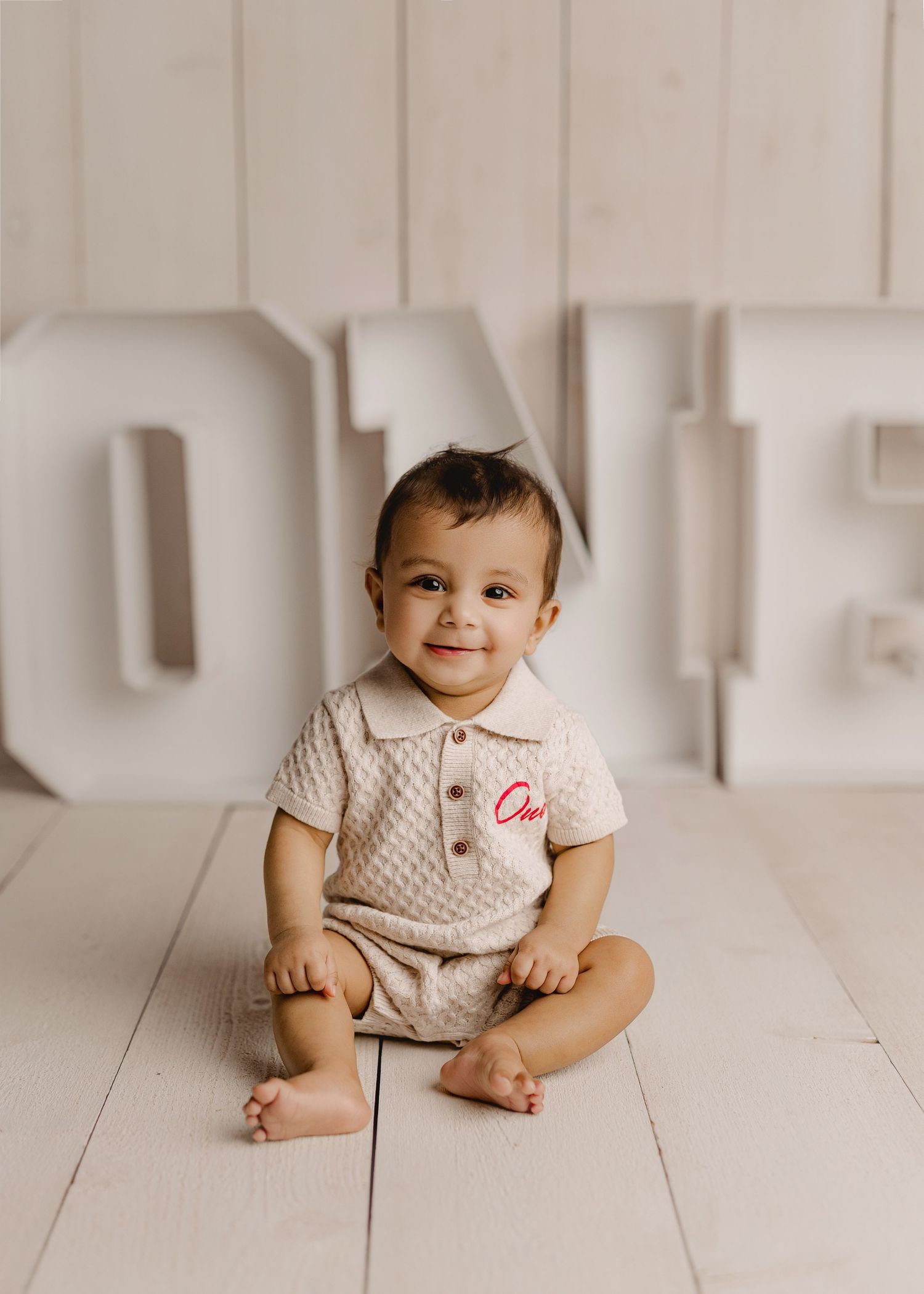 A smiling baby in a cream knit romper sits on a white wood floor in front of large ONE letters for a first birthday photo.