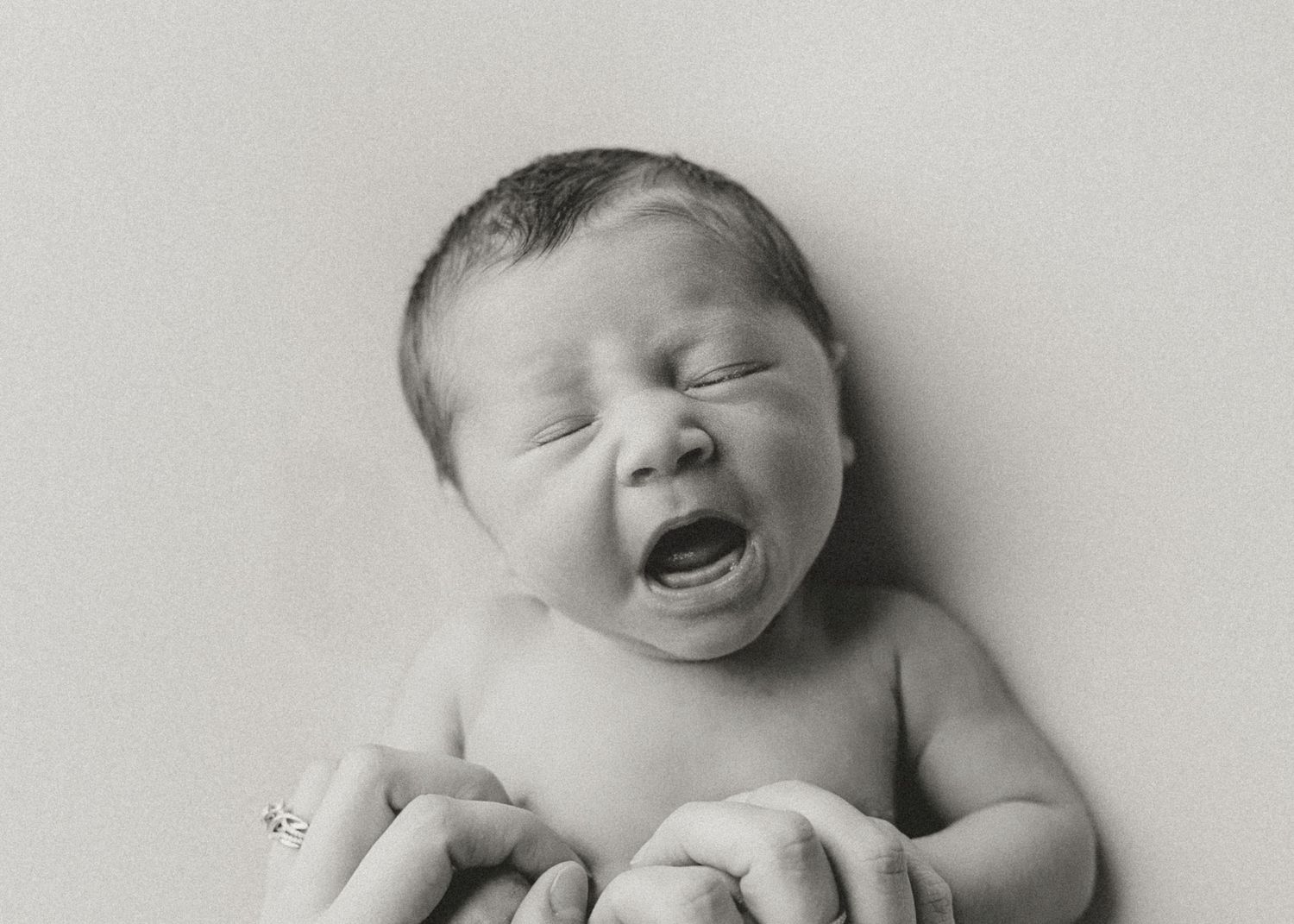 Black and white newborn photo of a crying baby being gently held by adult hands.