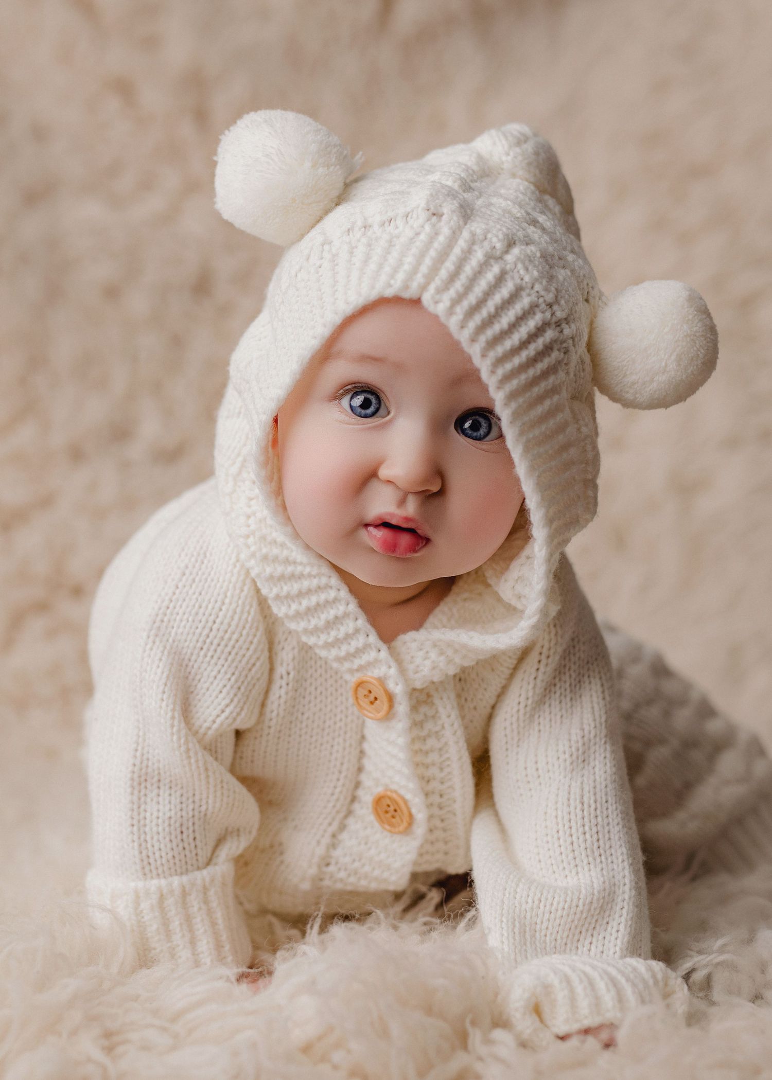 Baby in cream knit sweater and pom-pom hat crawling on white rug with blue eyes and tongue out. Southeast Michigan Milestone photographer.