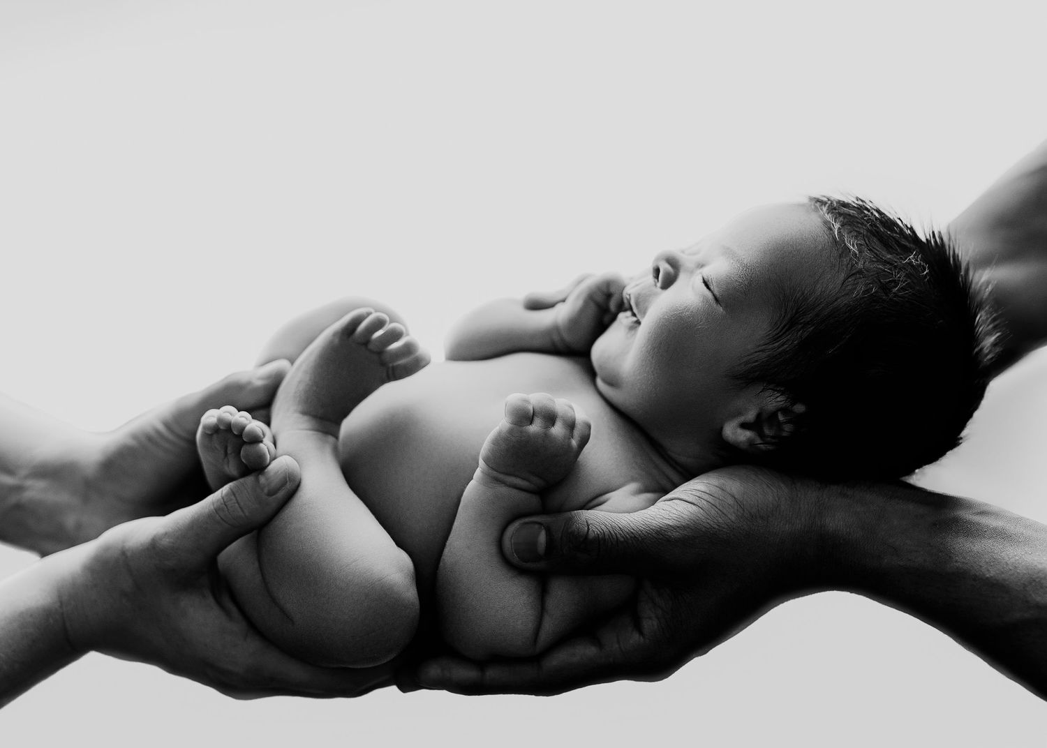 Black and white photo of a newborn baby cradled gently in adult hands against a white background.