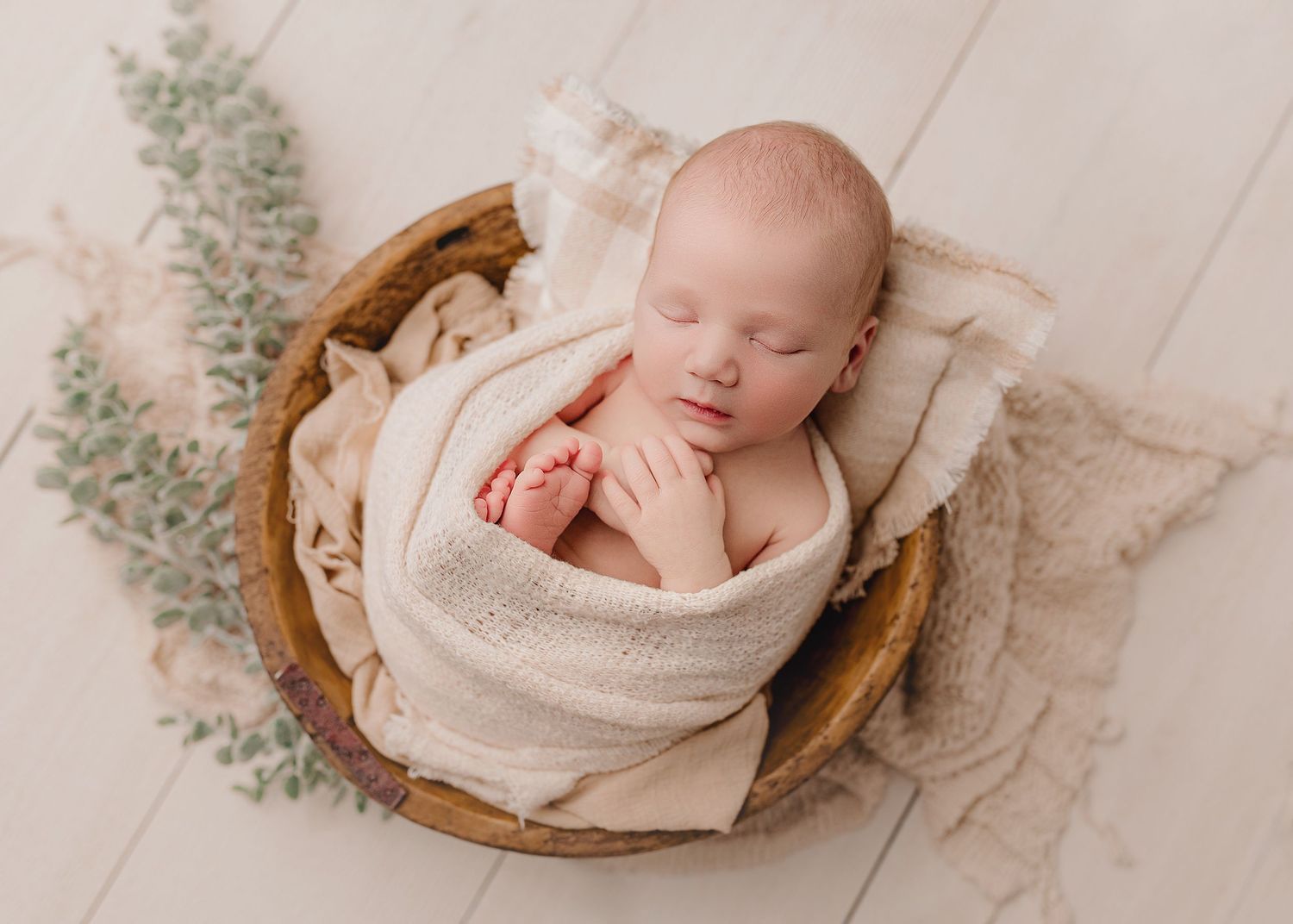 Sleeping newborn baby wrapped in cream swaddle cloth nestled in wooden bowl with soft lace and eucalyptus decor.