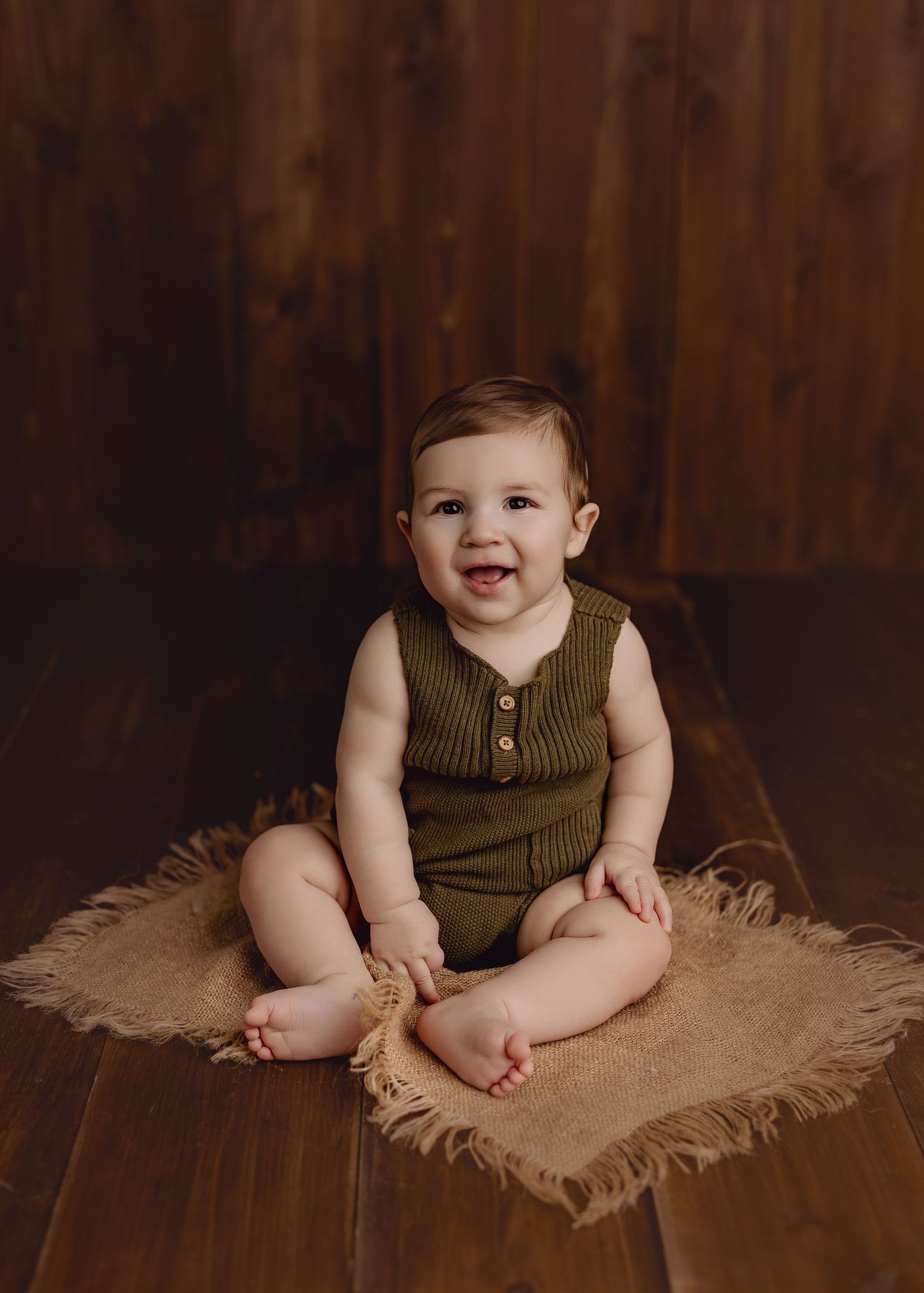 Smiling baby in olive green knit romper sitting on burlap rug against rustic wood backdrop.