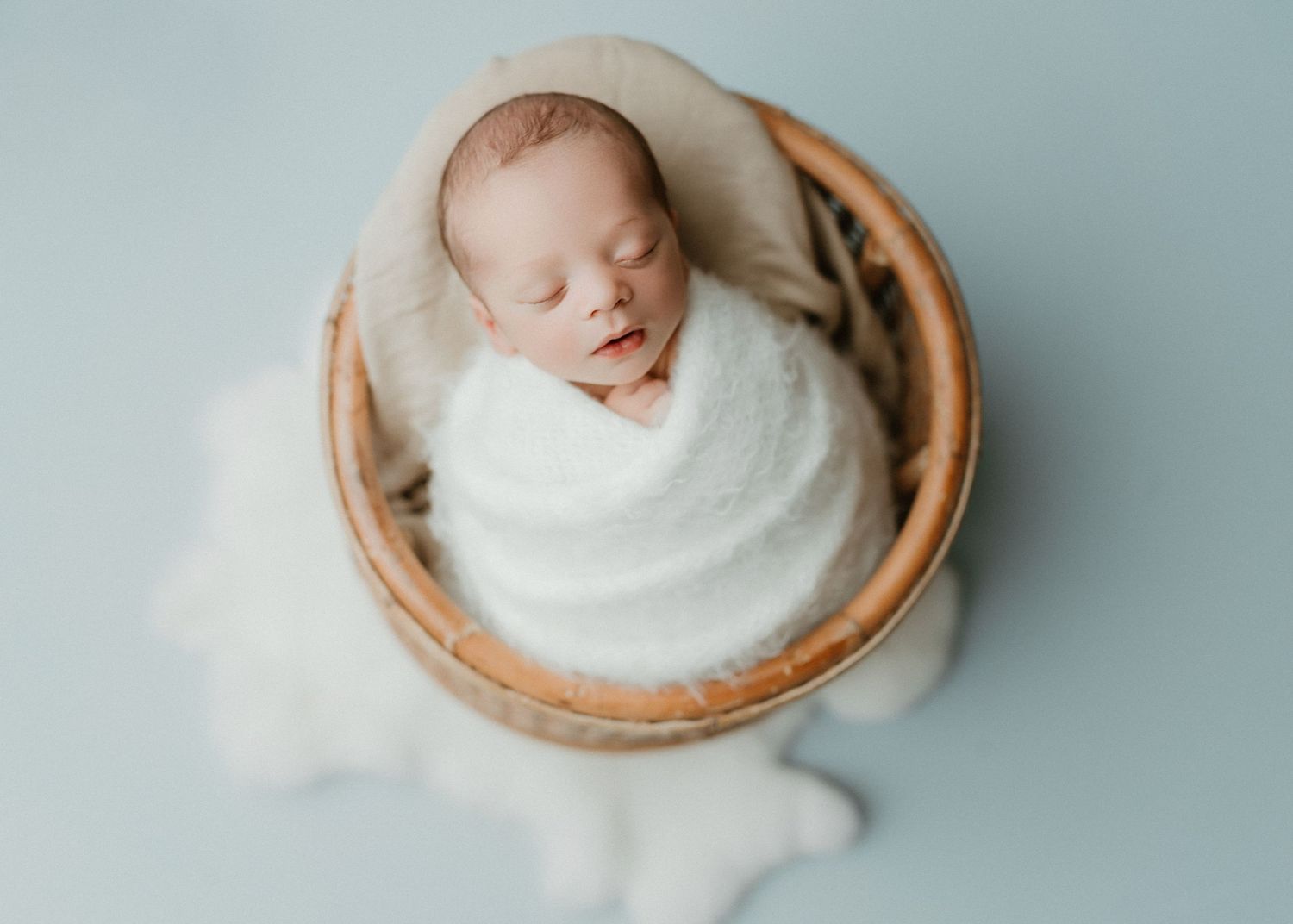 Sleeping newborn baby wrapped in white swaddle inside a wooden bowl on a soft blue background.