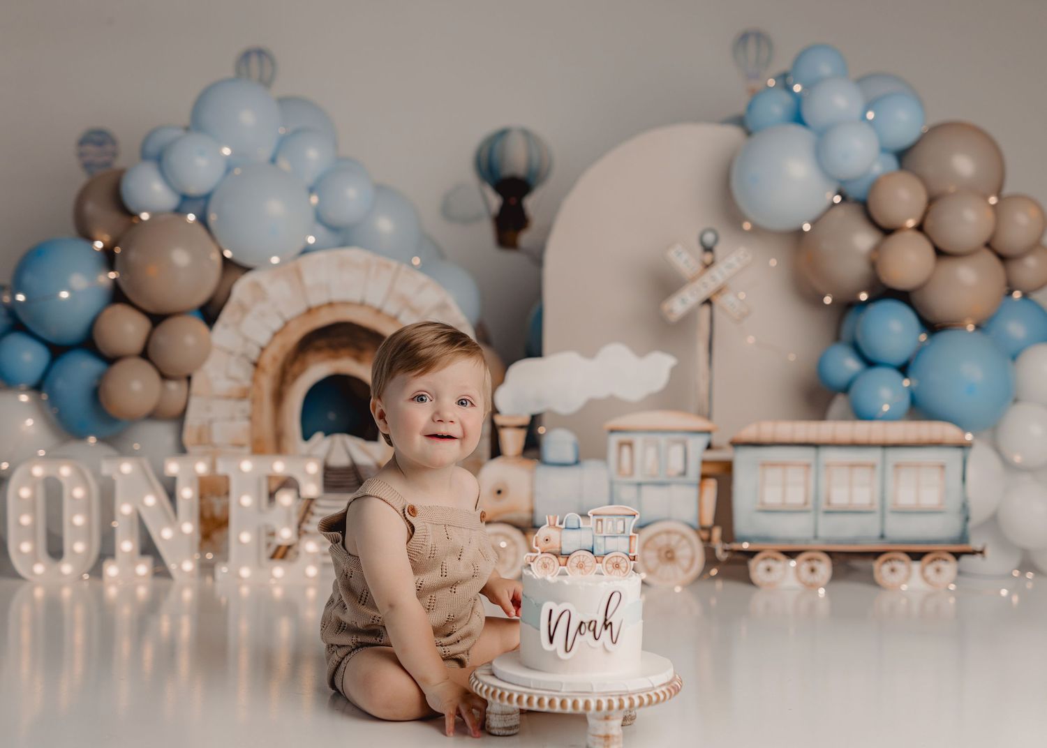 Baby boy sits by a cake at his first birthday party with blue and beige balloon decorations and a wooden train backdrop.