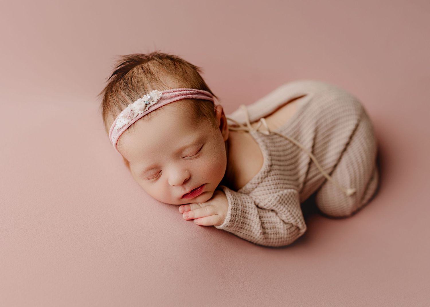 A newborn baby wearing a cream knit outfit lies peacefully sleeping on a soft pink backdrop.