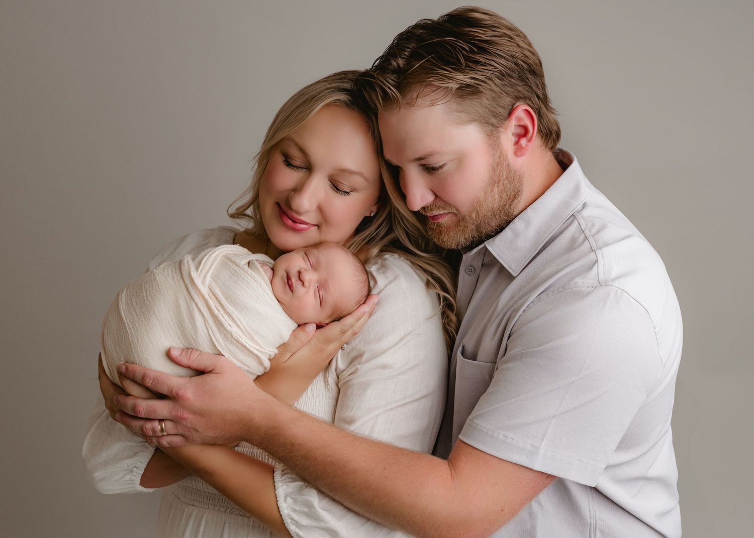 New parents tenderly hold their swaddled newborn baby in a soft, neutral-toned professional studio portrait.