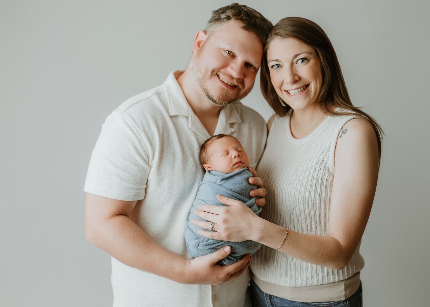 Smiling couple in white tops holding their swaddled newborn baby in a blue wrap against a light gray background.