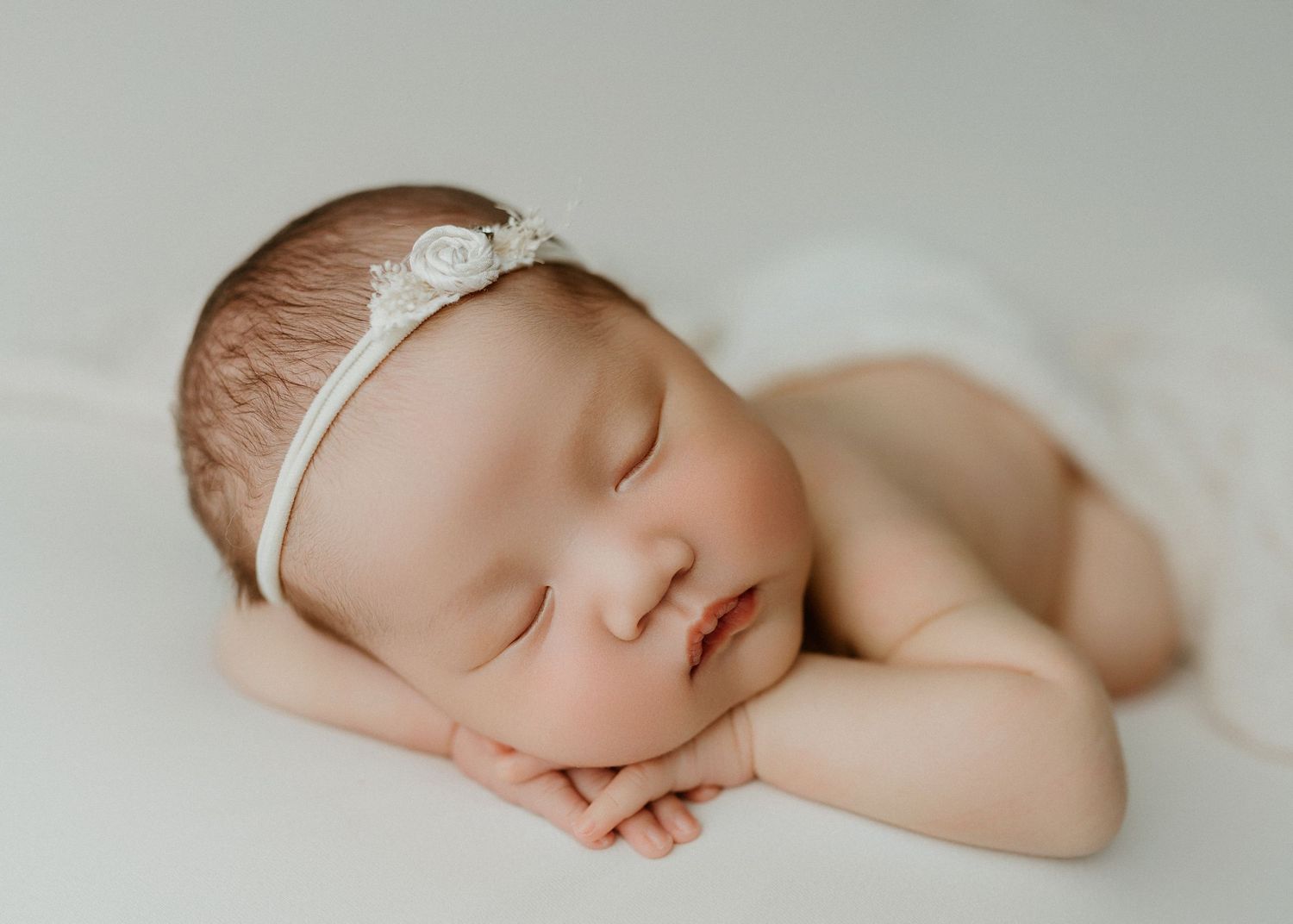 Peaceful newborn baby girl sleeping with hands under chin, wearing a delicate white floral headband on soft white backdrop.