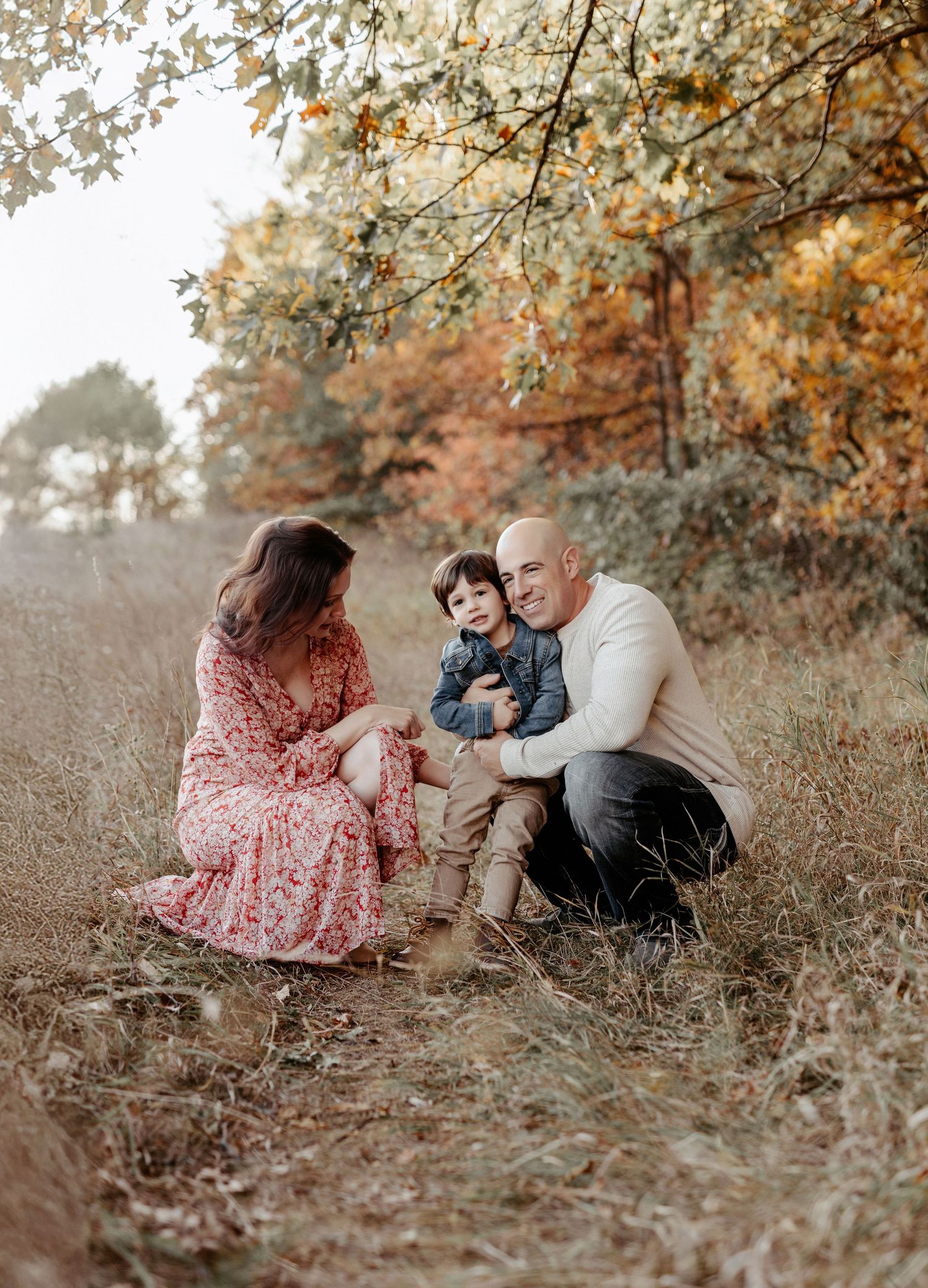 Family of three sitting in autumn field, parents smiling at toddler boy, surrounded by colorful fall foliage.