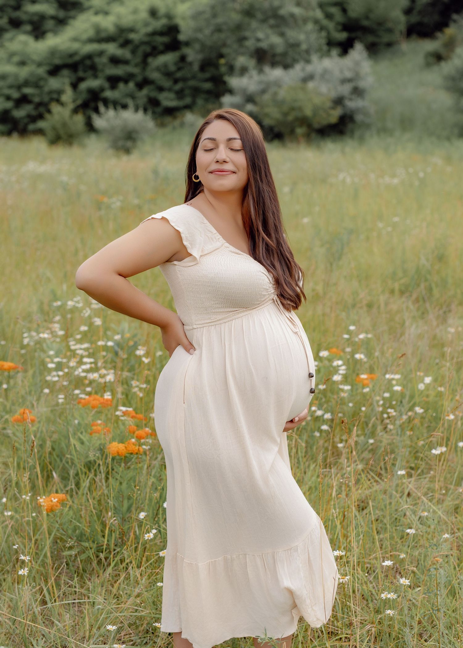 Pregnant woman in a cream flowy maternity dress cradling her baby bump in a sunlit meadow of wildflowers and daisies.