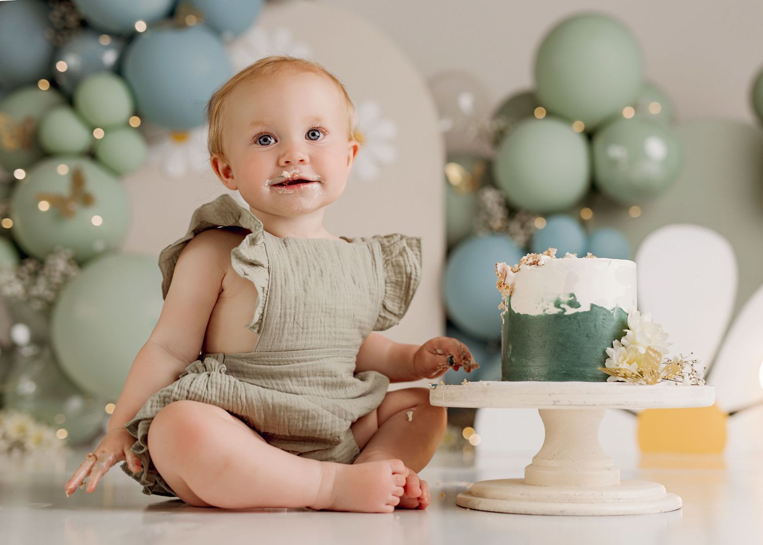 Baby girl in sage romper sits beside a green and white smash cake with balloon backdrop for first birthday photos.