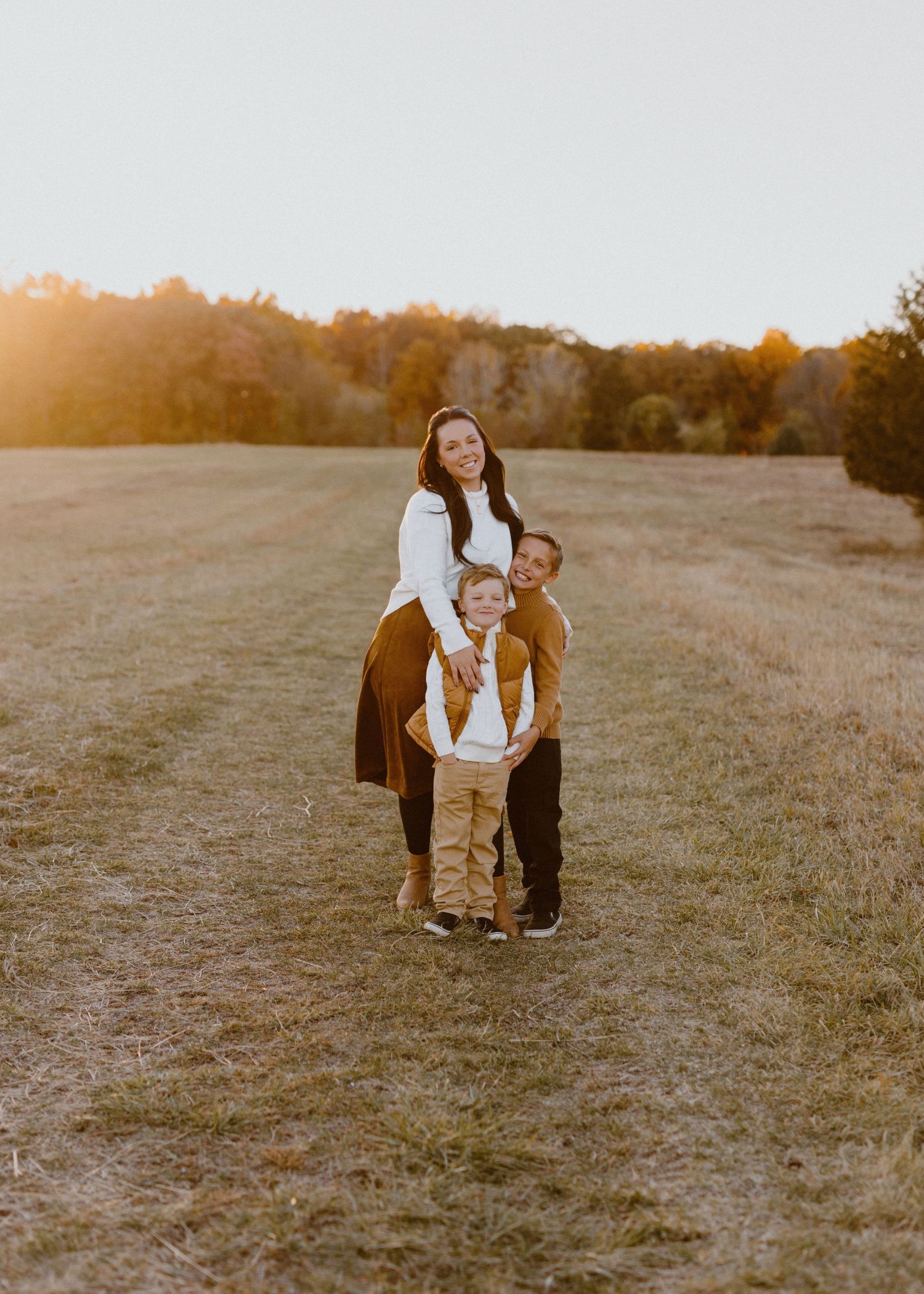 Family of three posing in golden hour sunlight in an open field with autumn trees in the background.