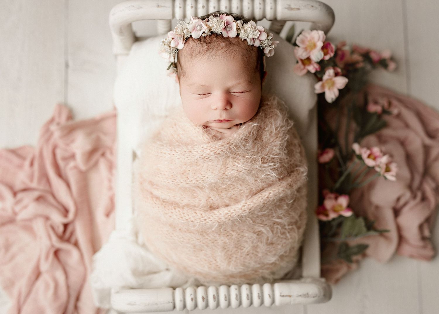 Newborn baby wrapped in cream lace swaddle with floral crown, sleeping peacefully in a white vintage mini bed.