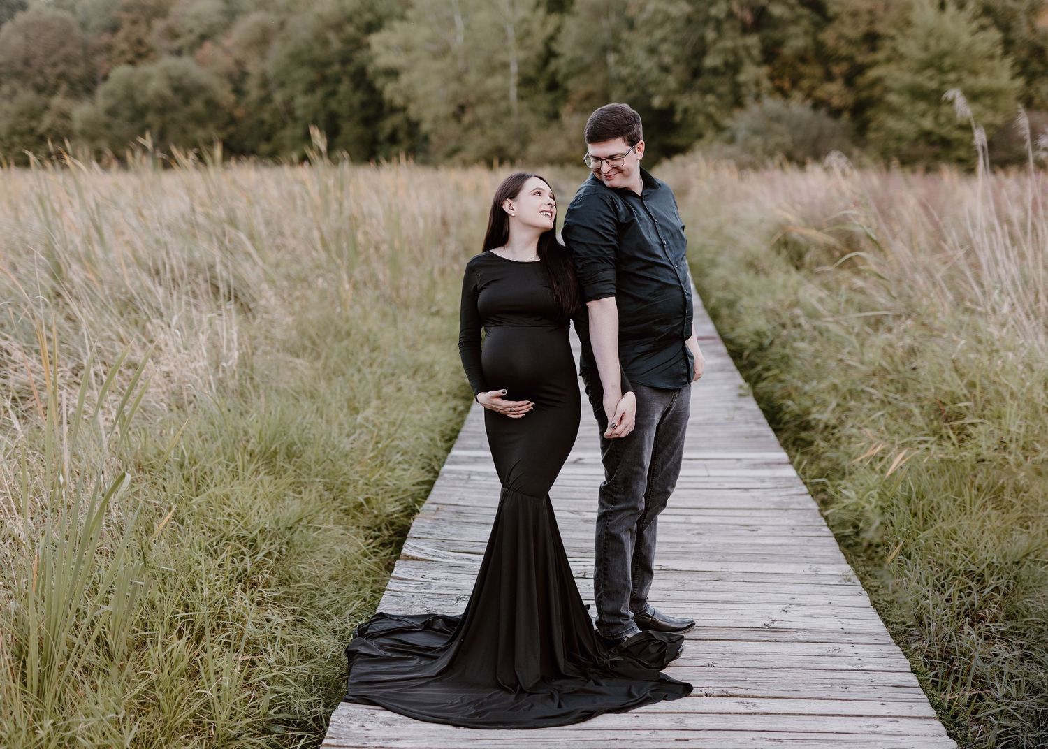 Expecting couple holds hands on a wooden boardwalk through tall grass, the woman in a black fitted maternity gown with a flowing train.
