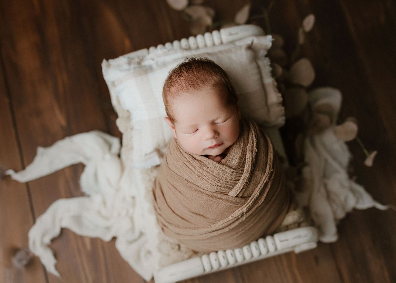 Newborn baby wrapped in tan swaddle sleeping peacefully on white mini bed with pillow on wood floor.
