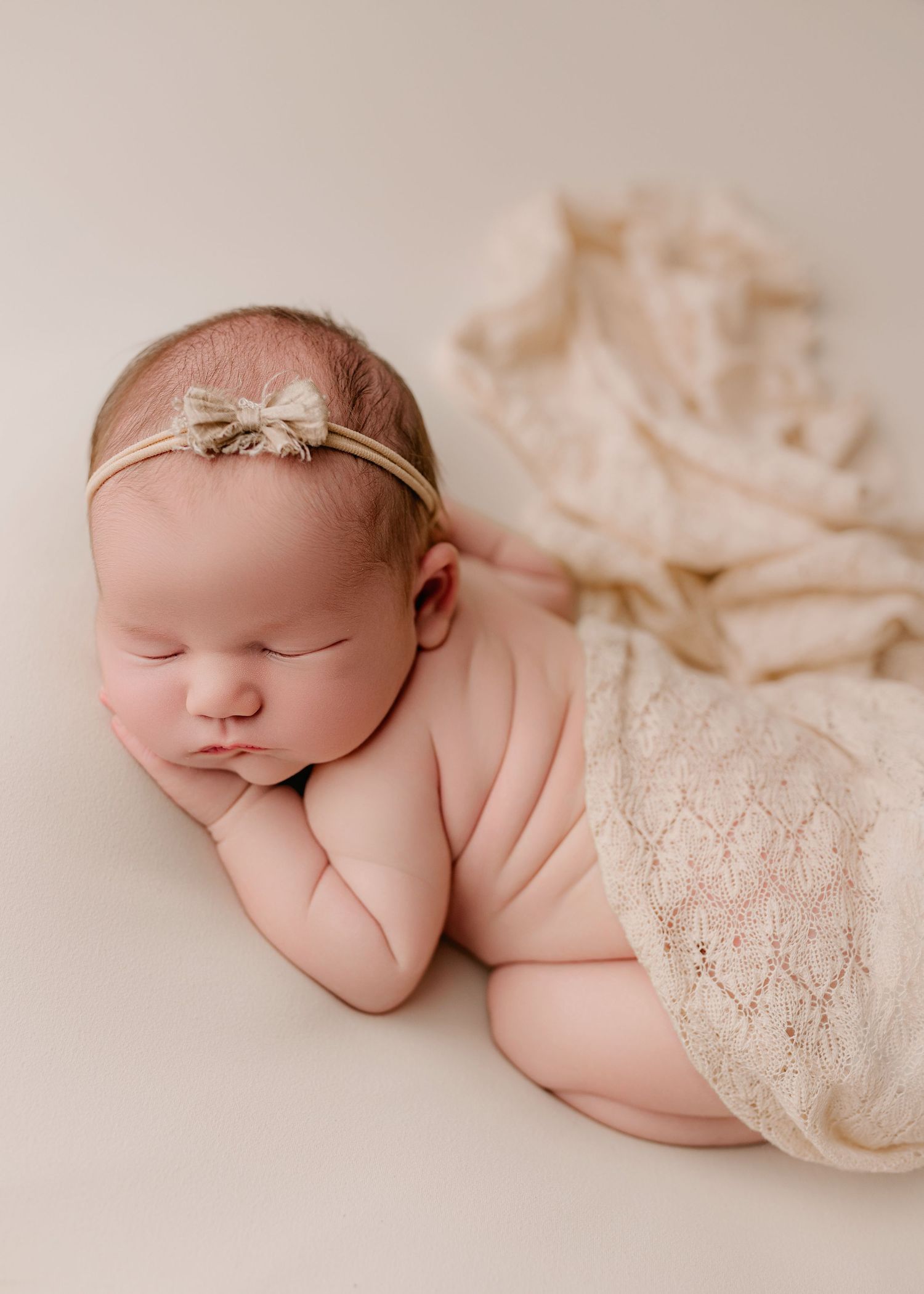 Sleeping newborn wrapped in delicate lace blanket lying on cream colored fabric in artistic photography series.